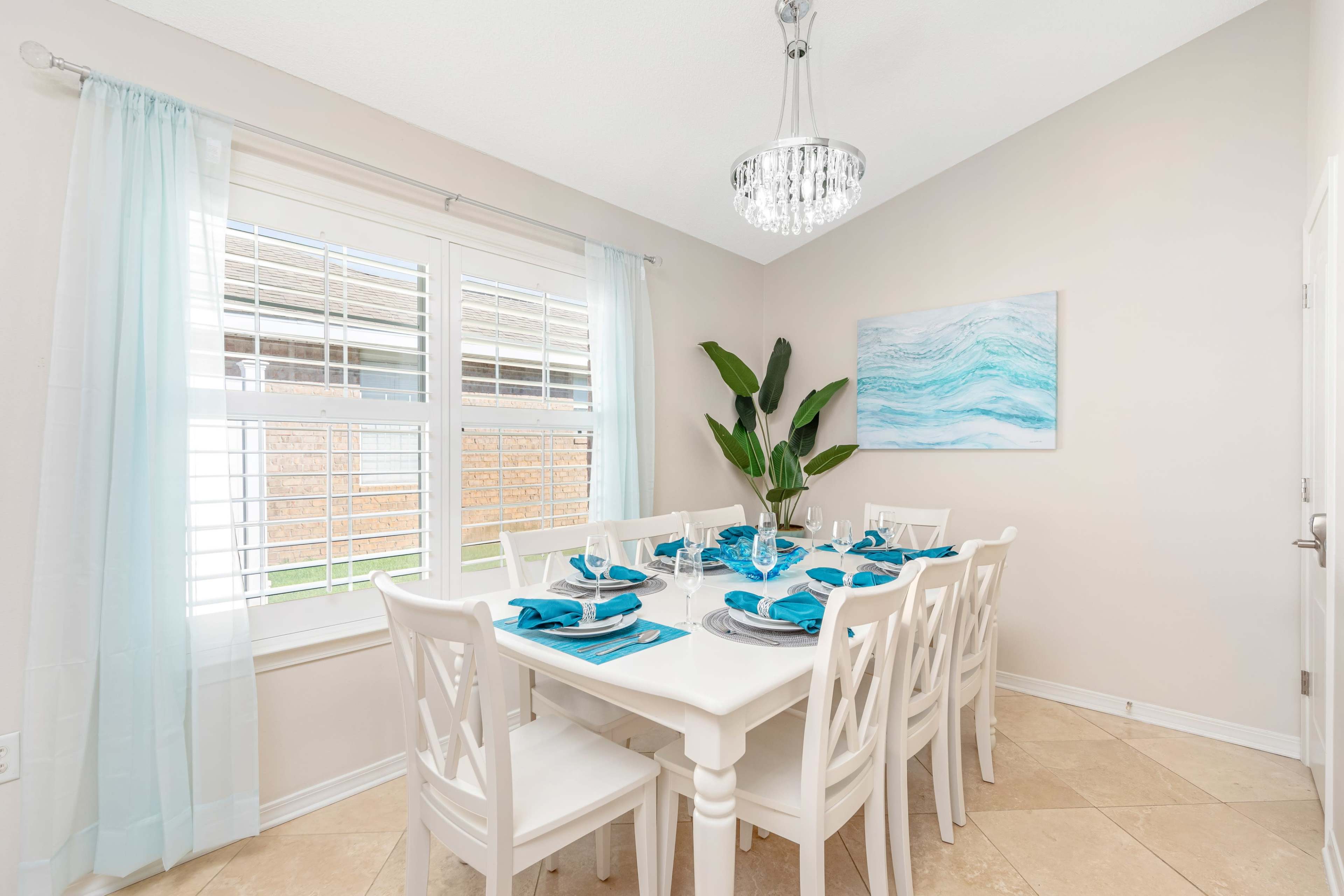 A dining area features a long white table set with turquoise dinnerware, surrounded by white chairs, and a large painting of ocean waves on the wall.