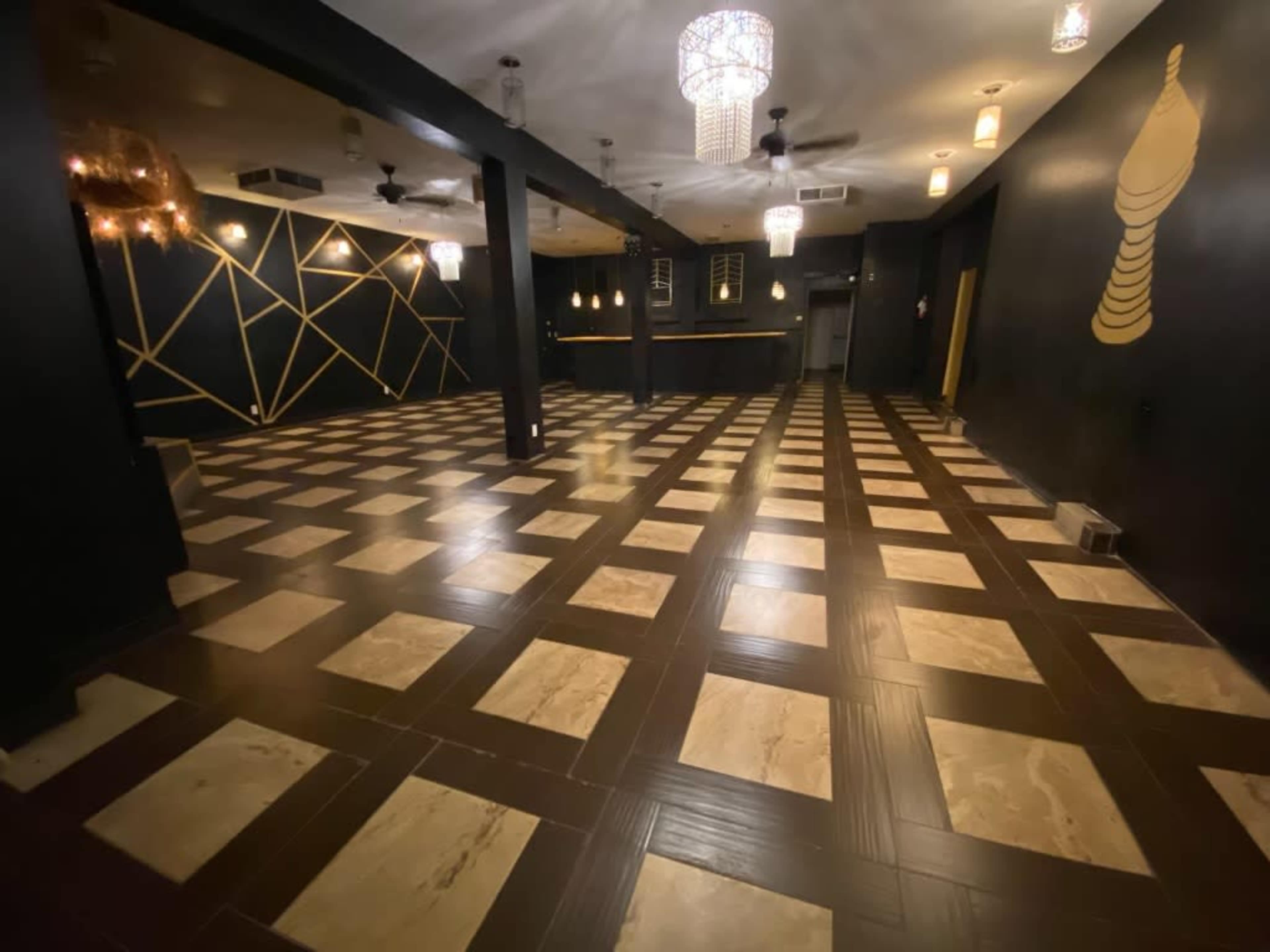 The image shows a spacious indoor area with a patterned floor featuring alternating light and dark tiles, accented by modern lighting fixtures hanging from the ceiling.