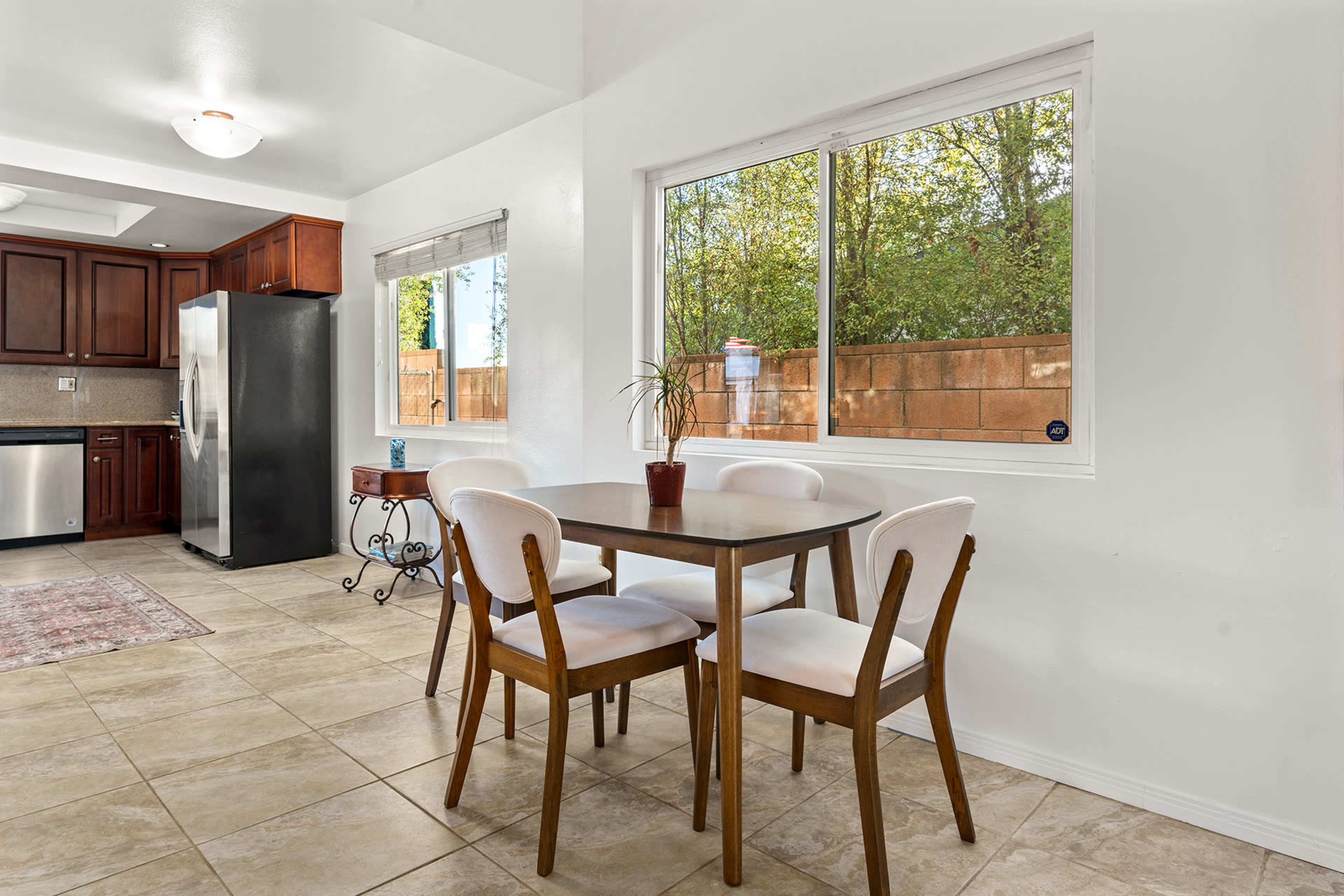 A small dining area features a table with four chairs, illuminated by natural light from a window overlooking a green outdoor space.