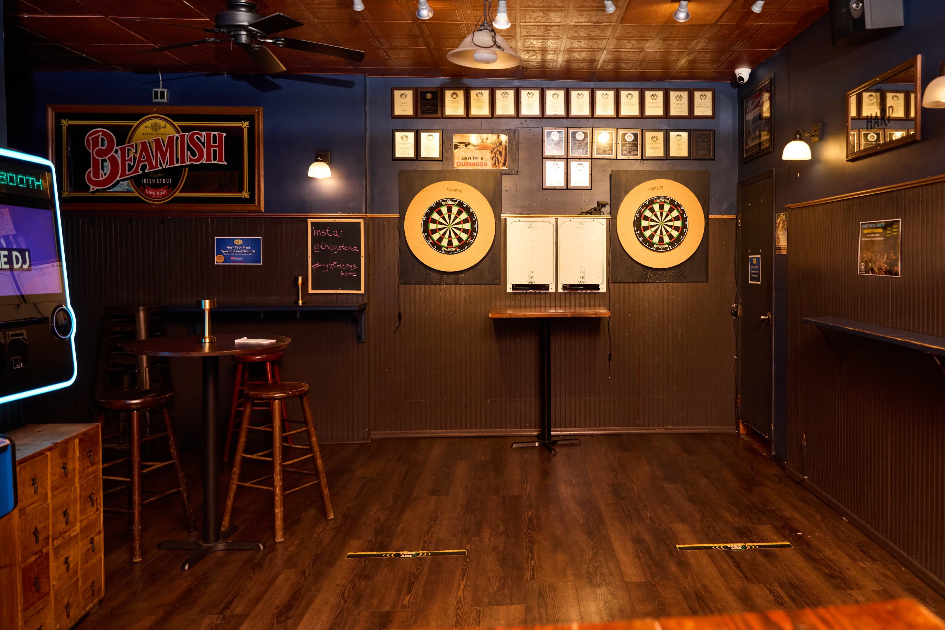 The image shows a dimly lit bar area with two dartboards on the wall, surrounded by plaques, and a few tables and chairs placed around.