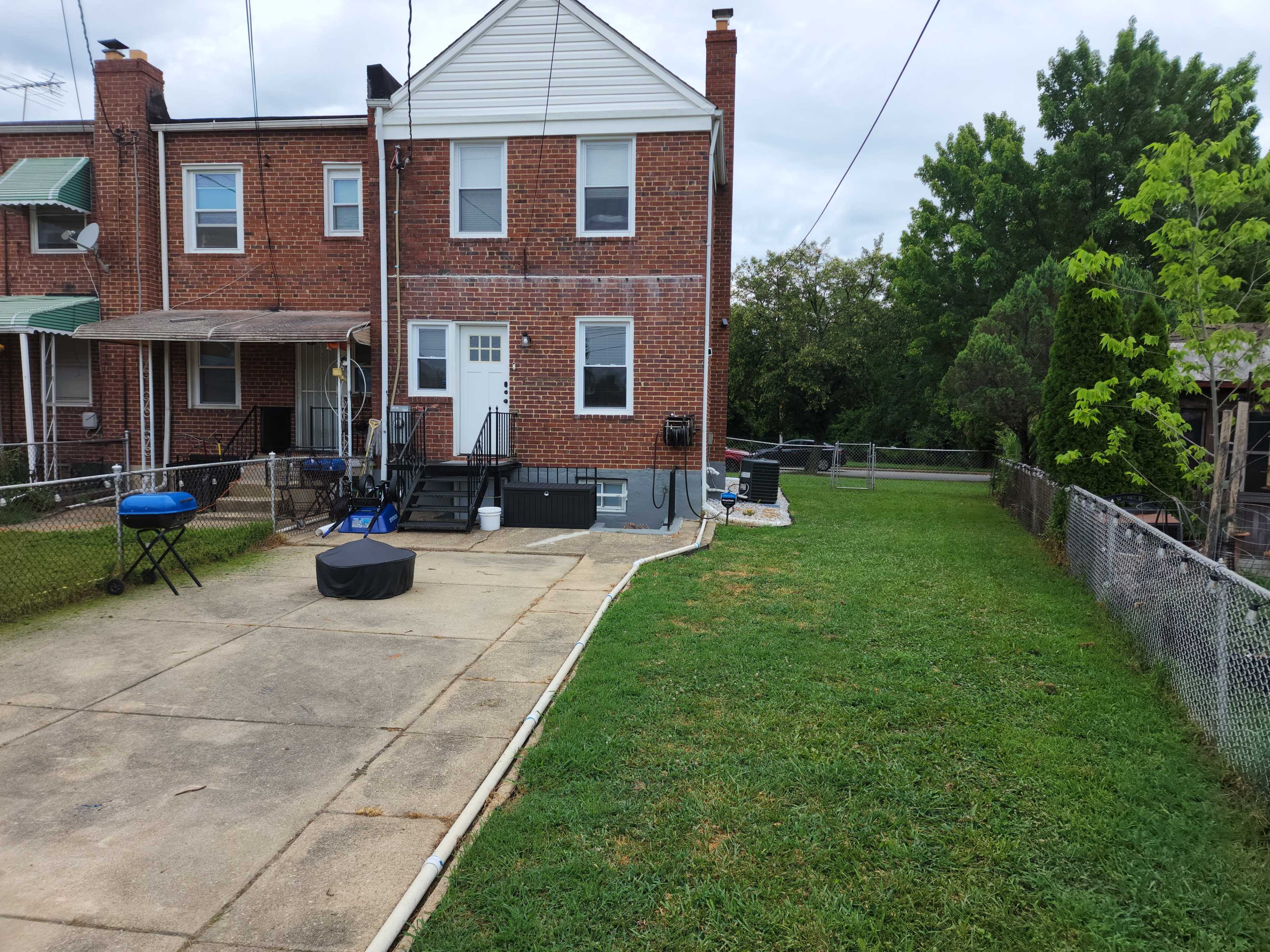 The image shows the back of a brick townhouse with a paved area and a grassy yard, surrounded by a fence.
