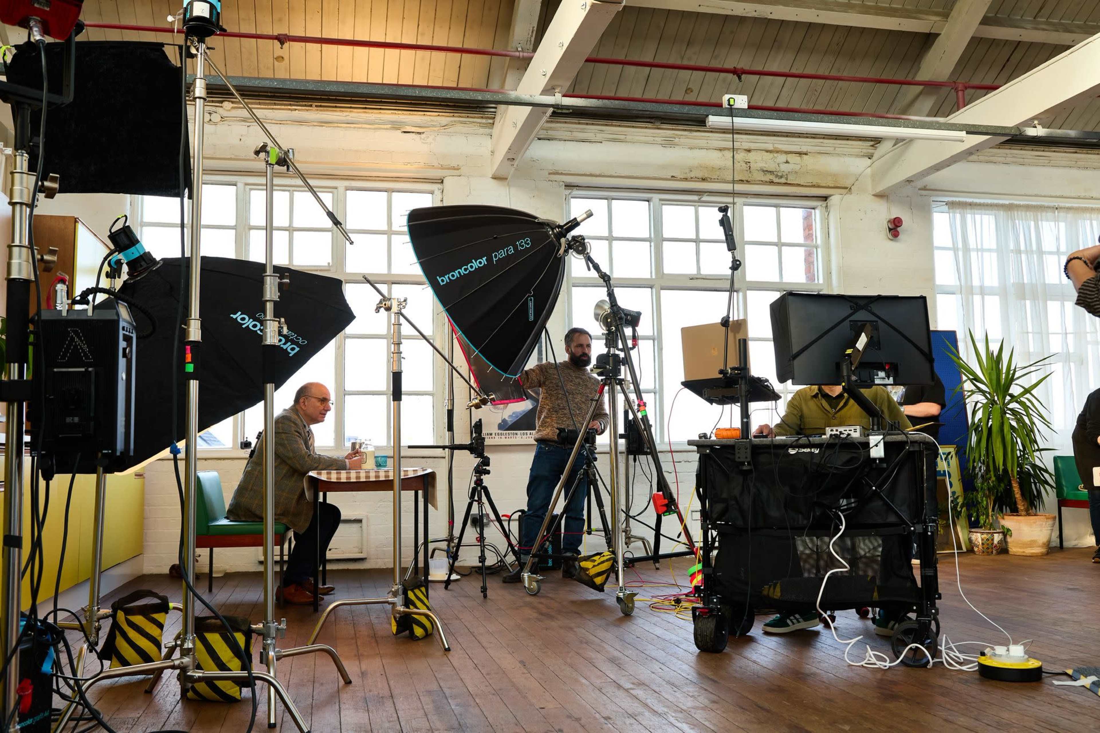 A photography studio is set up with multiple lights, cameras, and equipment, while a man sits at a table and another person adjusts the setup.
