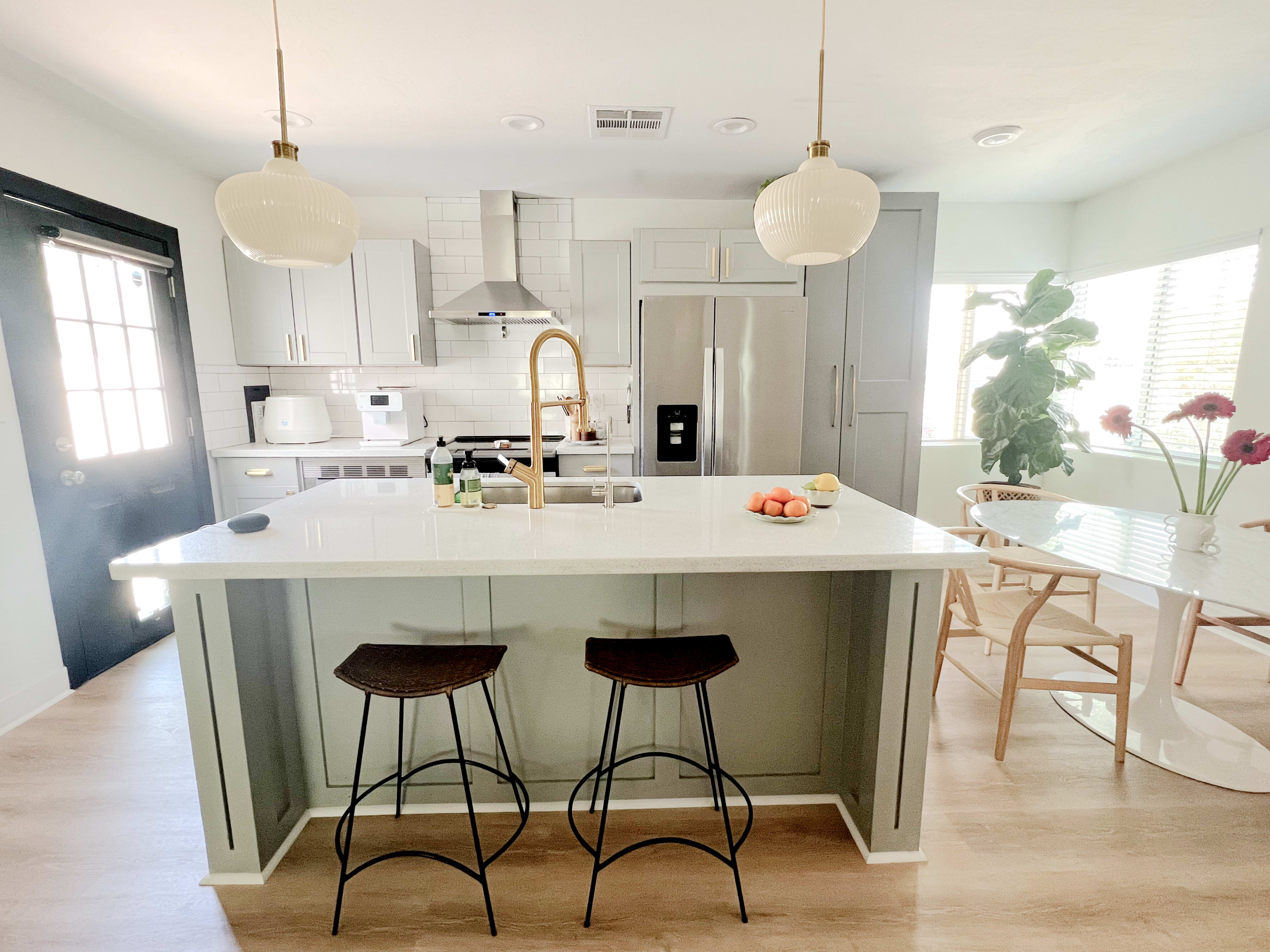 A modern kitchen with a large island, two bar stools, stainless steel appliances, and a dining table next to a window with plants and fruit.