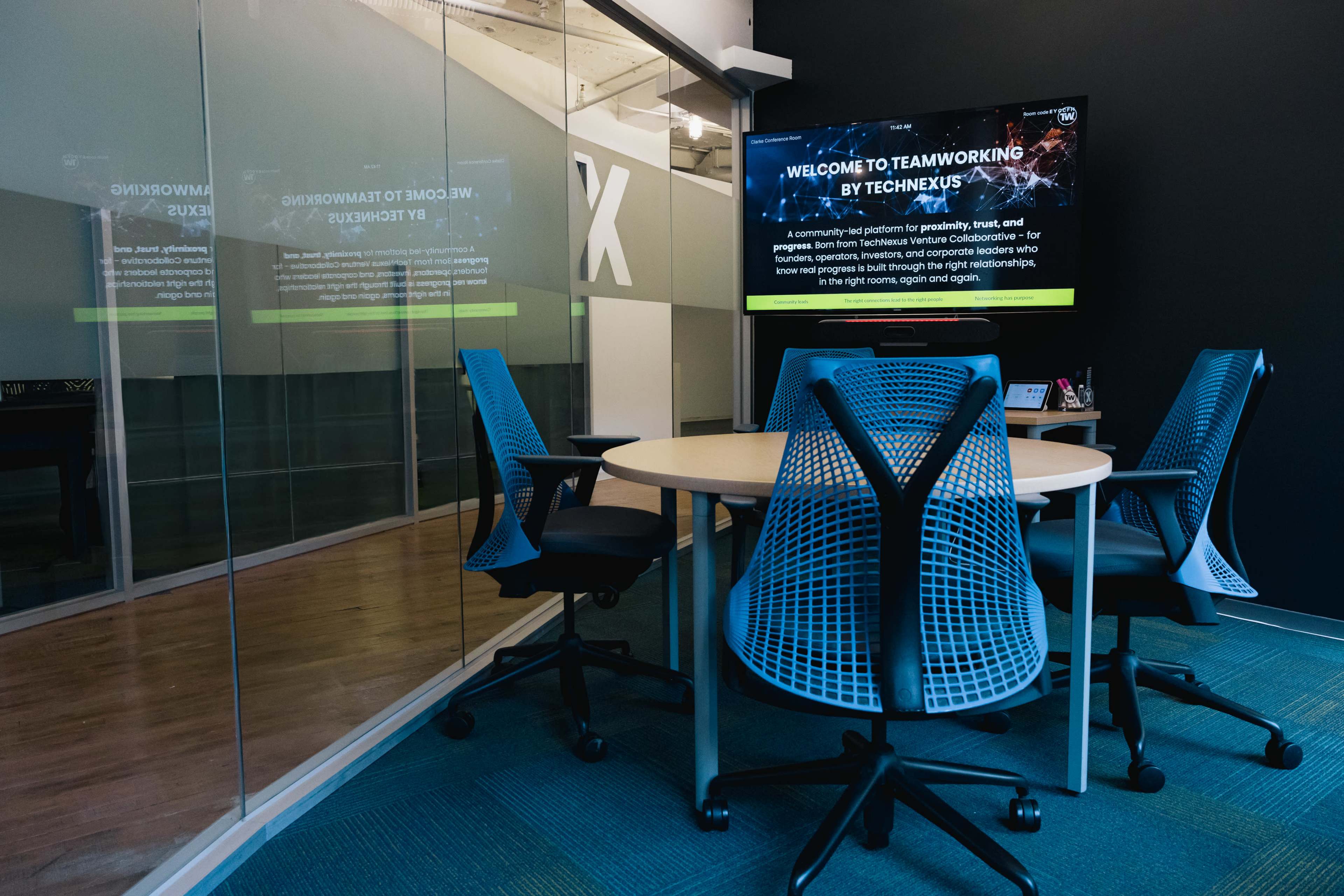 A conference room with a round table and four ergonomic chairs is shown, featuring a large screen displaying a welcome message on one wall and glass panels separating it from the adjoining space.