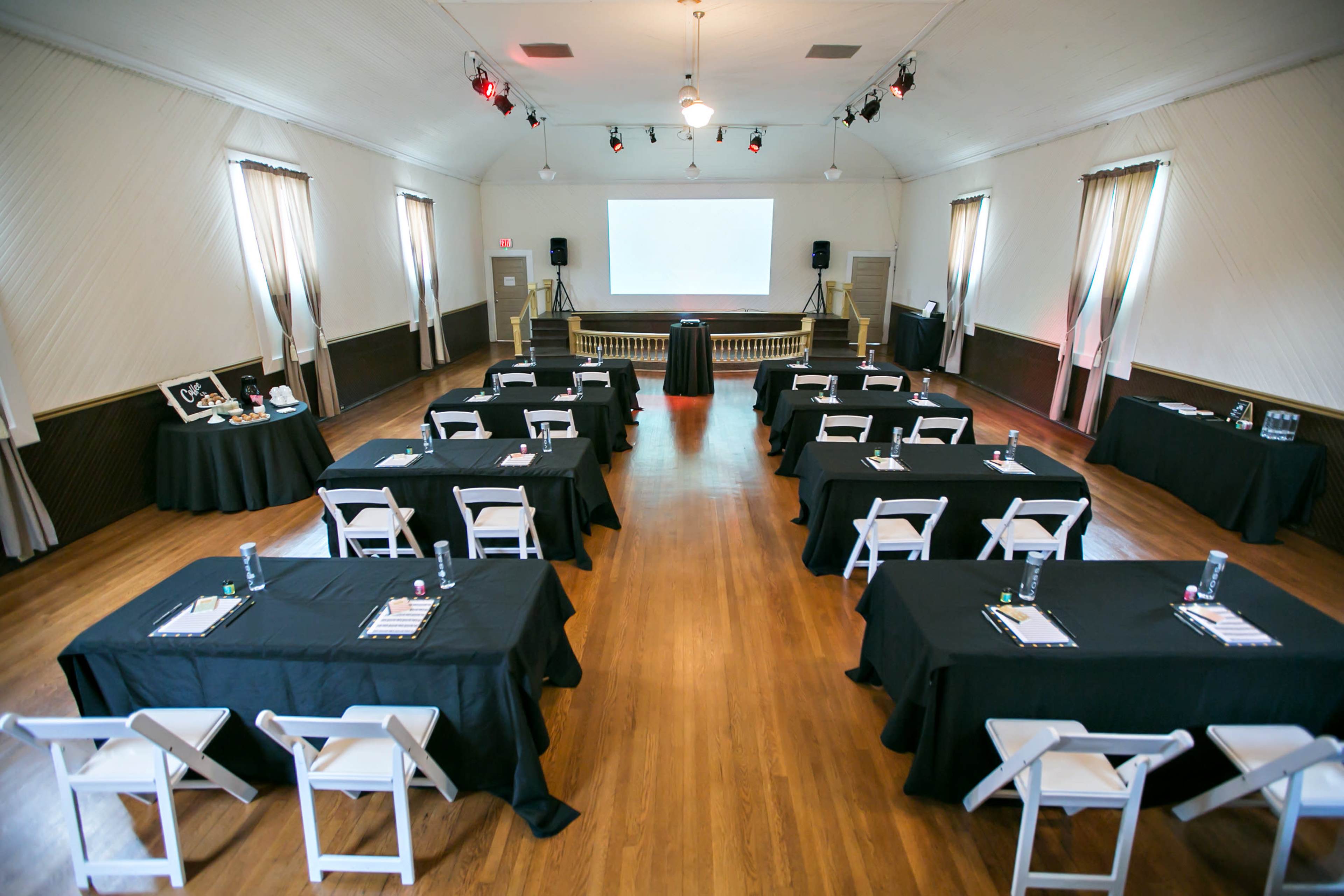 A spacious conference room set up with several tables arranged in a U-shape, each covered with black cloth and accompanied by chairs, facing a central presentation area with a projector screen.