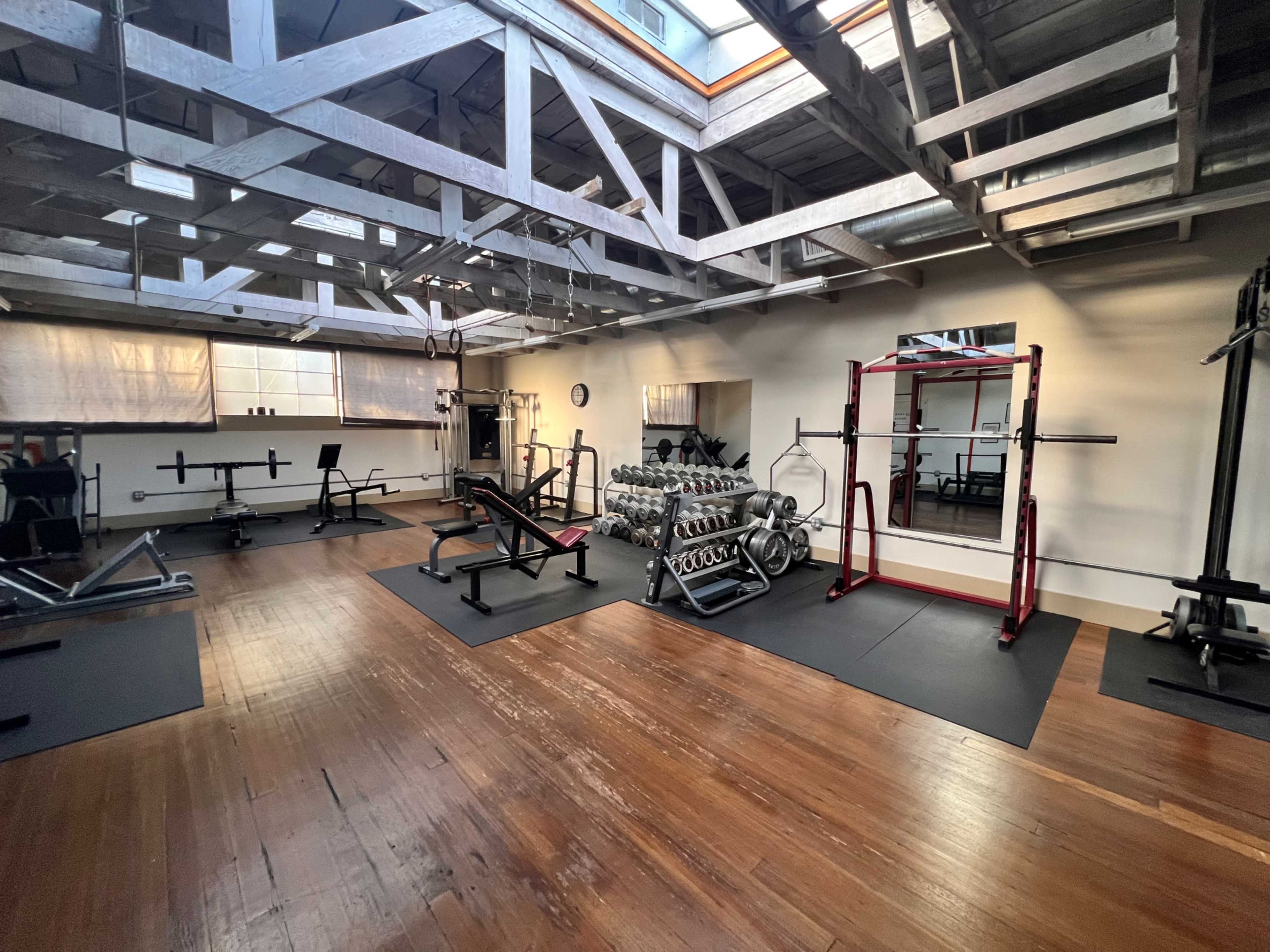 A gym with various weightlifting equipment, benches, and a rack of dumbbells on a wooden floor beneath a ceiling supported by wooden beams.