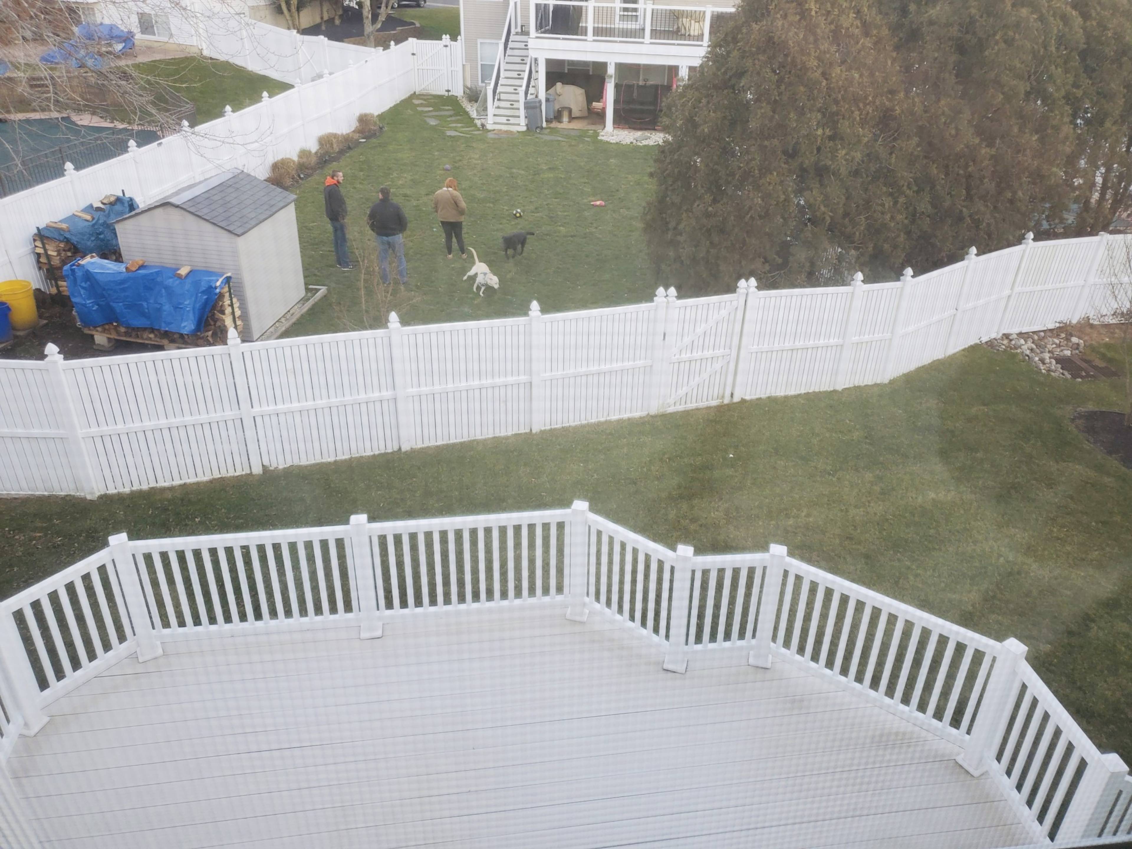 A group of three people and two dogs are in a fenced backyard with a white vinyl fence and a deck in the foreground.