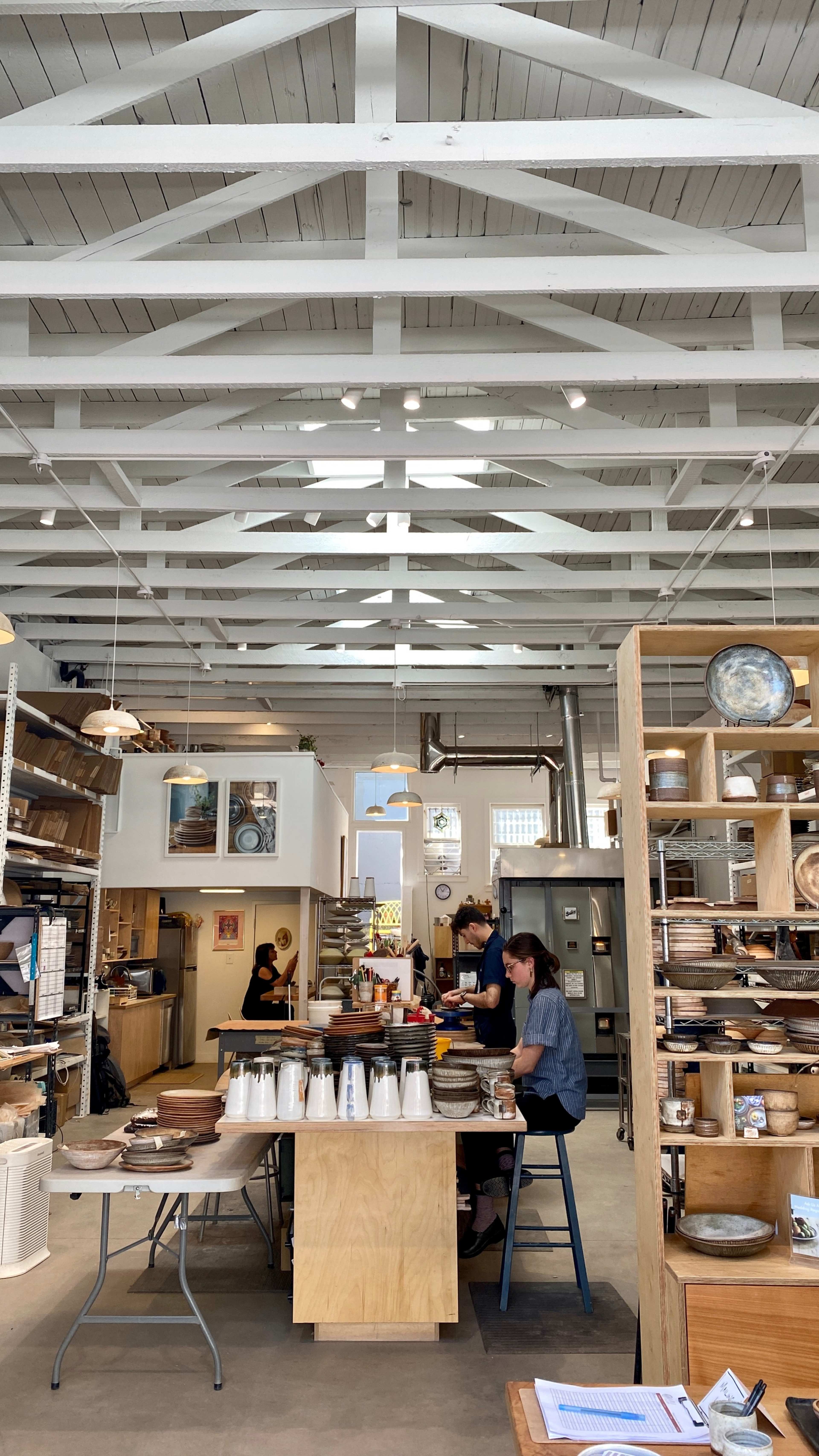 A bright, open café features wooden shelves filled with plates and utensils, while two customers engage at a central table.