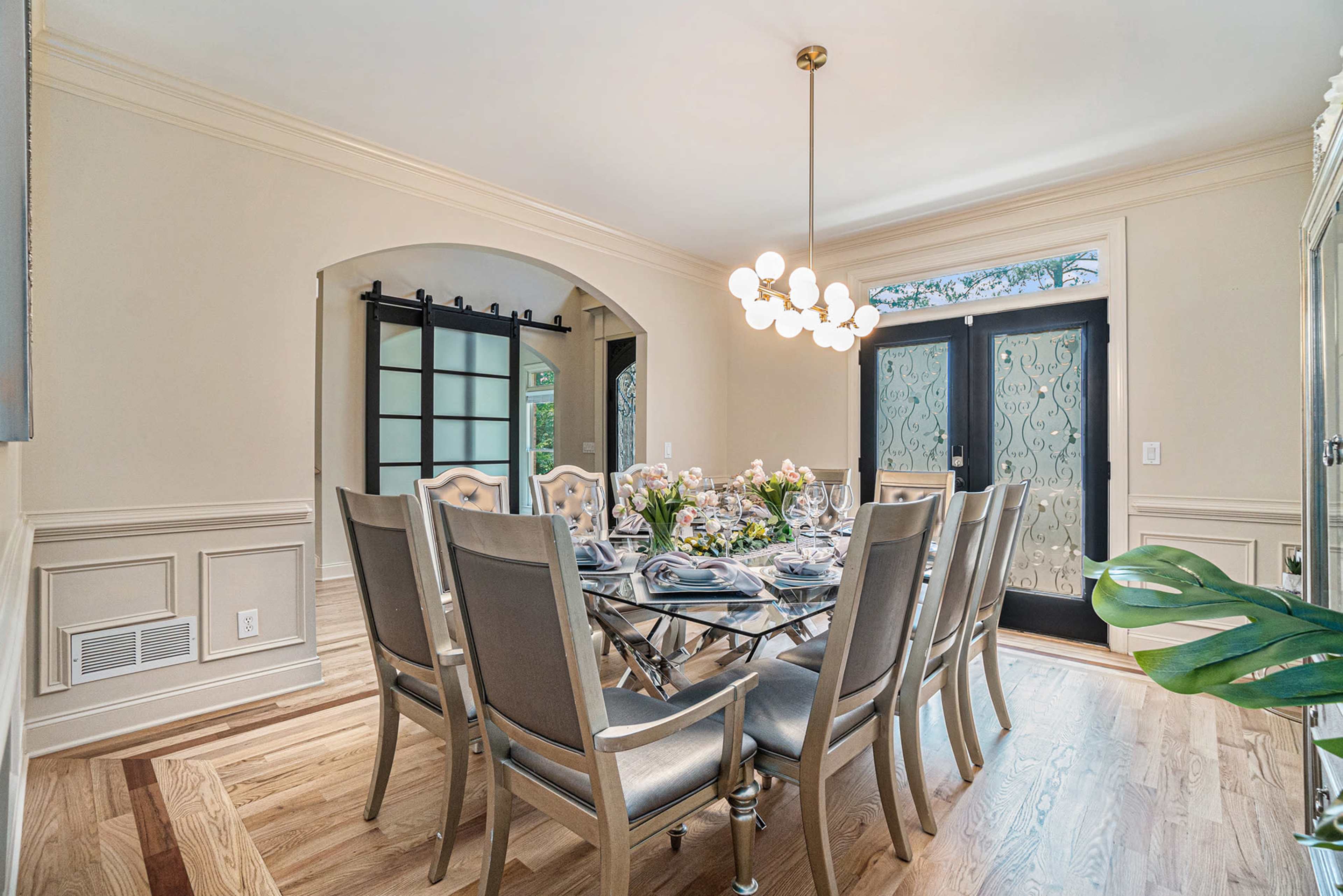 A spacious dining room features a long table set for a meal, with elegant chairs, modern light fixtures, and large glass doors leading to an outdoor area.
