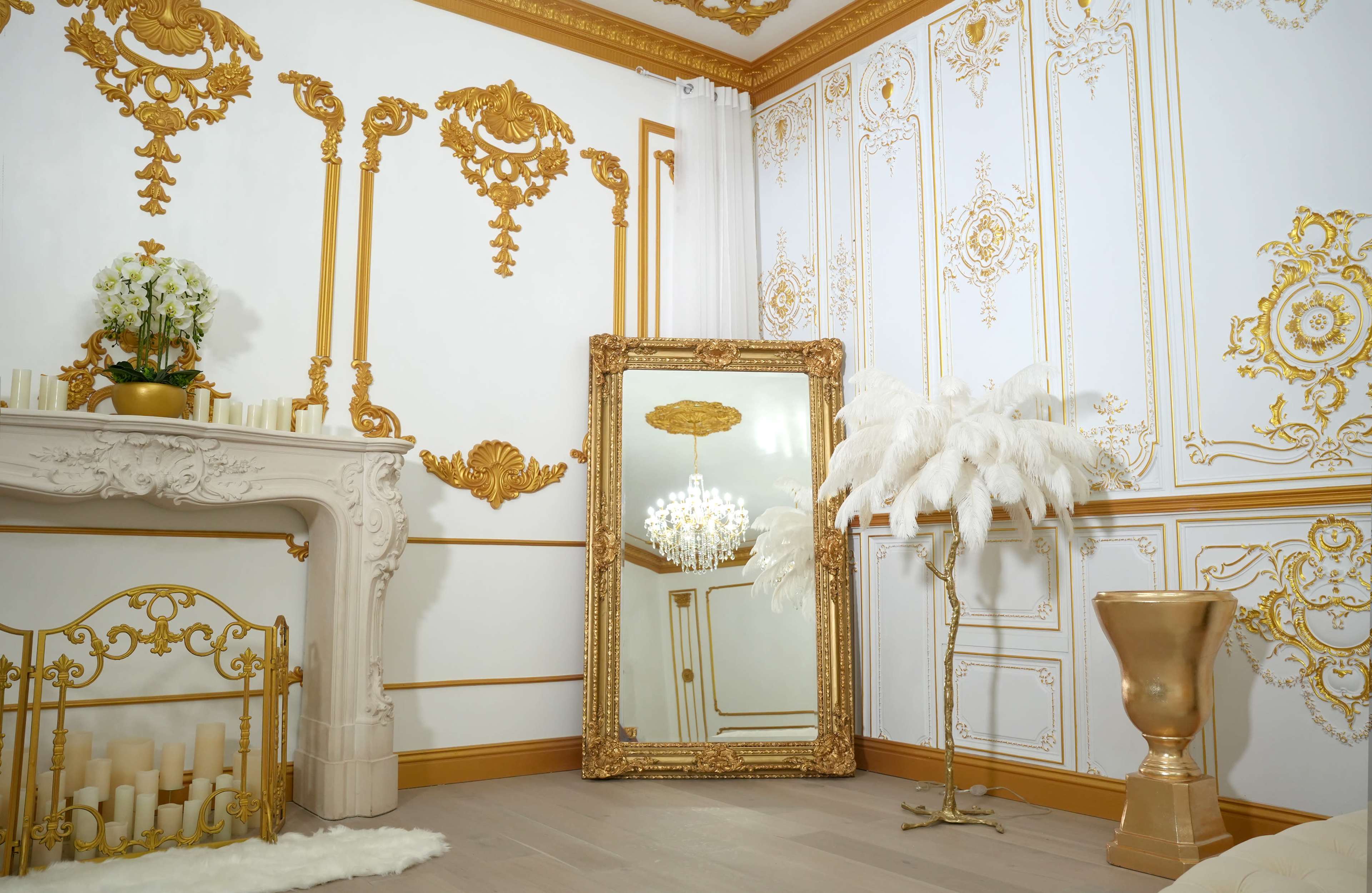 An elegantly decorated room featuring ornate gold accents, a large mirror, a chandelier, and decorative elements such as a fireplace and tall white feathers.