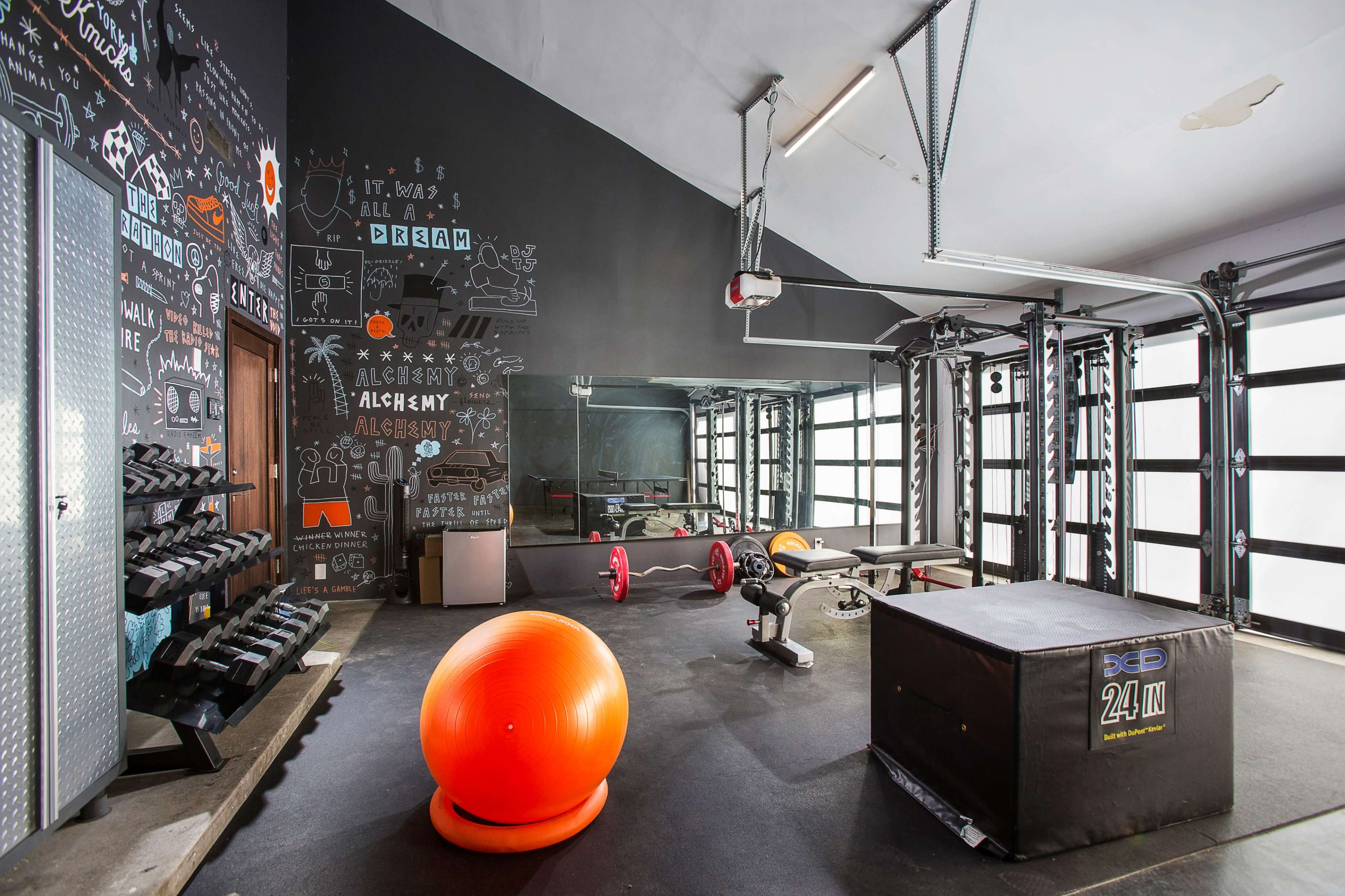 A fitness gym equipped with dumbbells, resistance machines, a bench, a stability ball, and a plyometric box, all set against a wall with motivational chalkboard art.