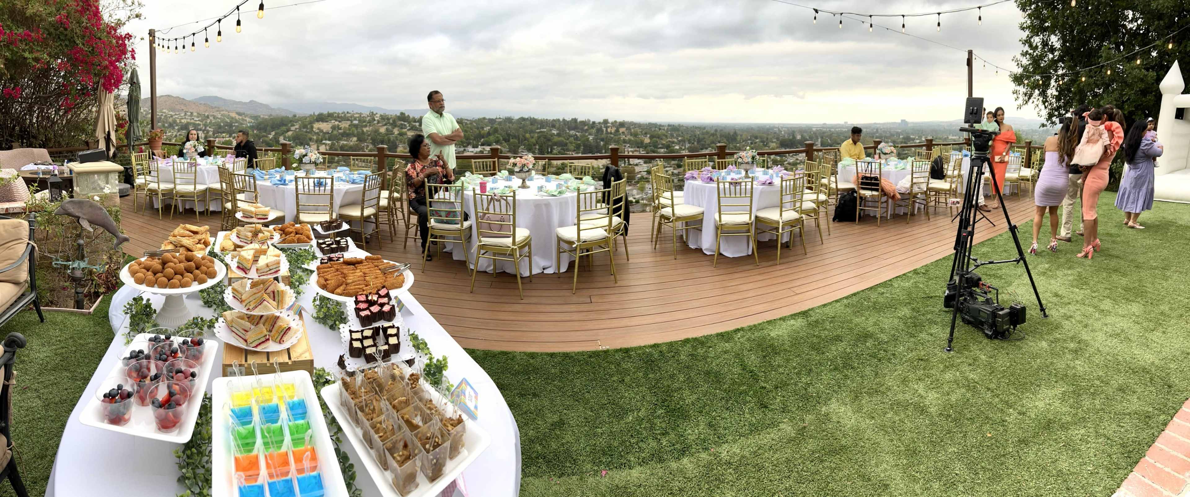 The outdoor venue features neatly arranged tables with white linens and various desserts displayed on a table, overlooking a scenic view.