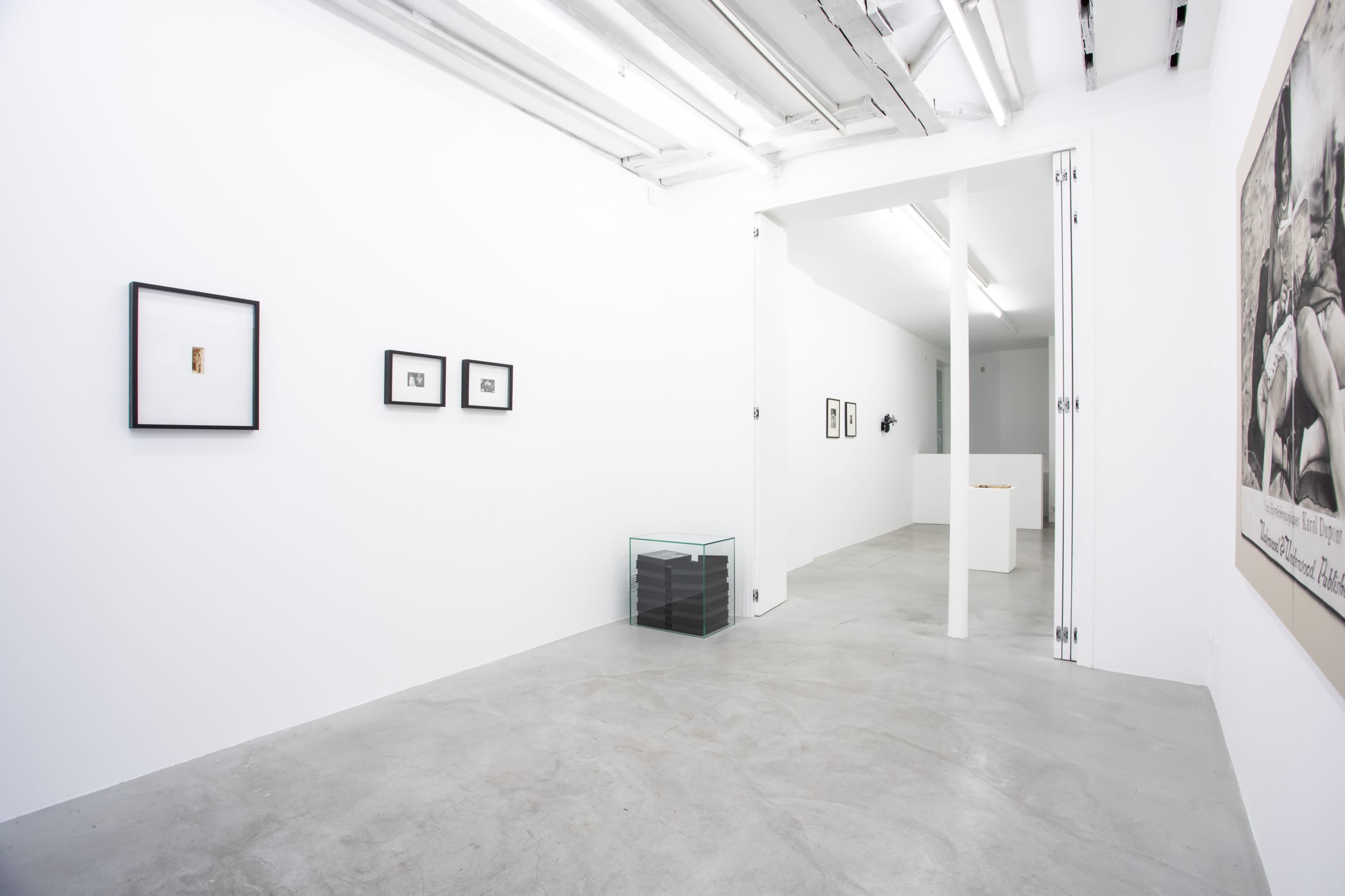 The image shows a minimalist art gallery with white walls, featuring several framed artworks and a glass display case.