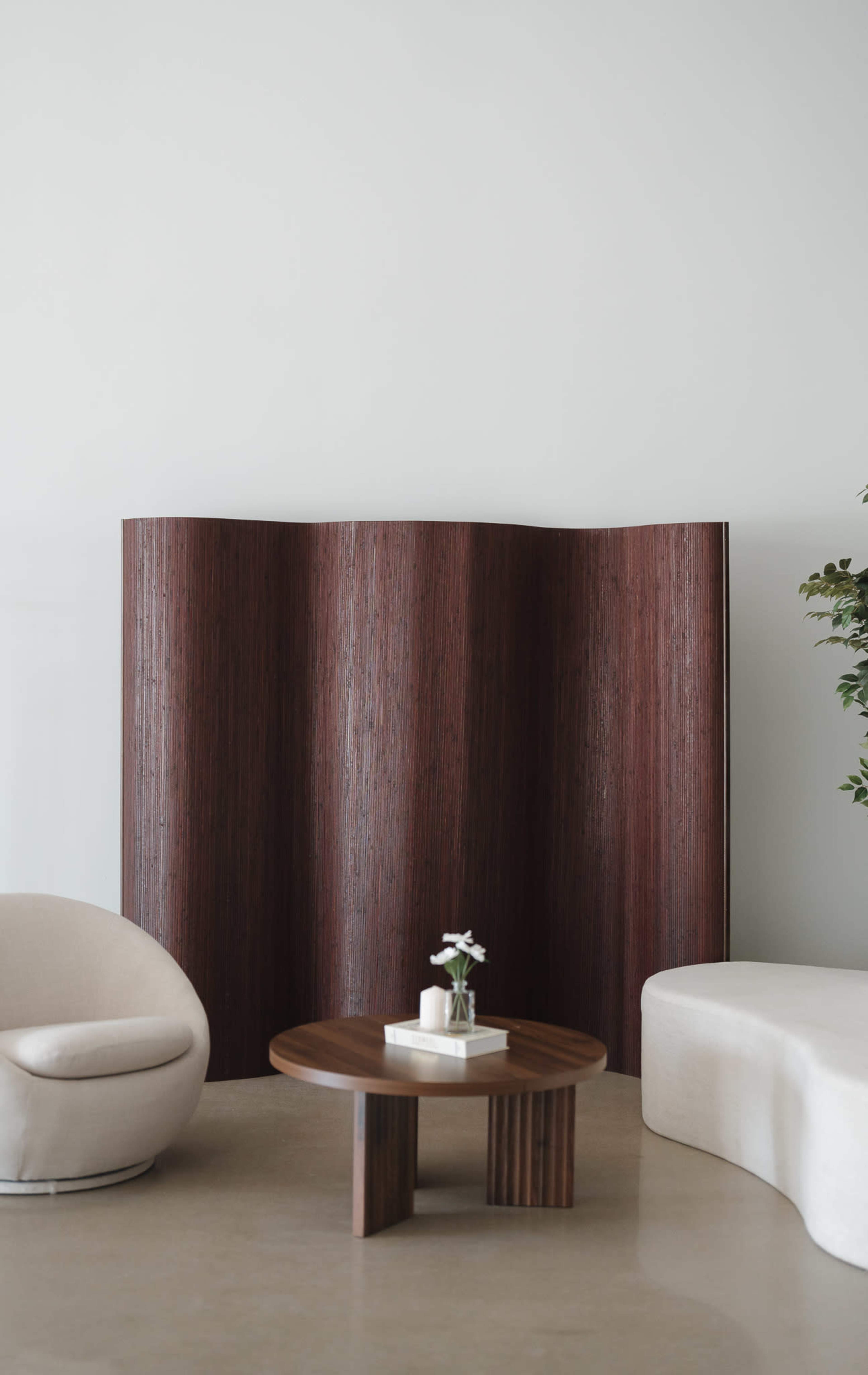 A modern interior scene features a curved wooden divider, a round table with a vase of flowers, and a minimalist chair in a neutral-toned space.