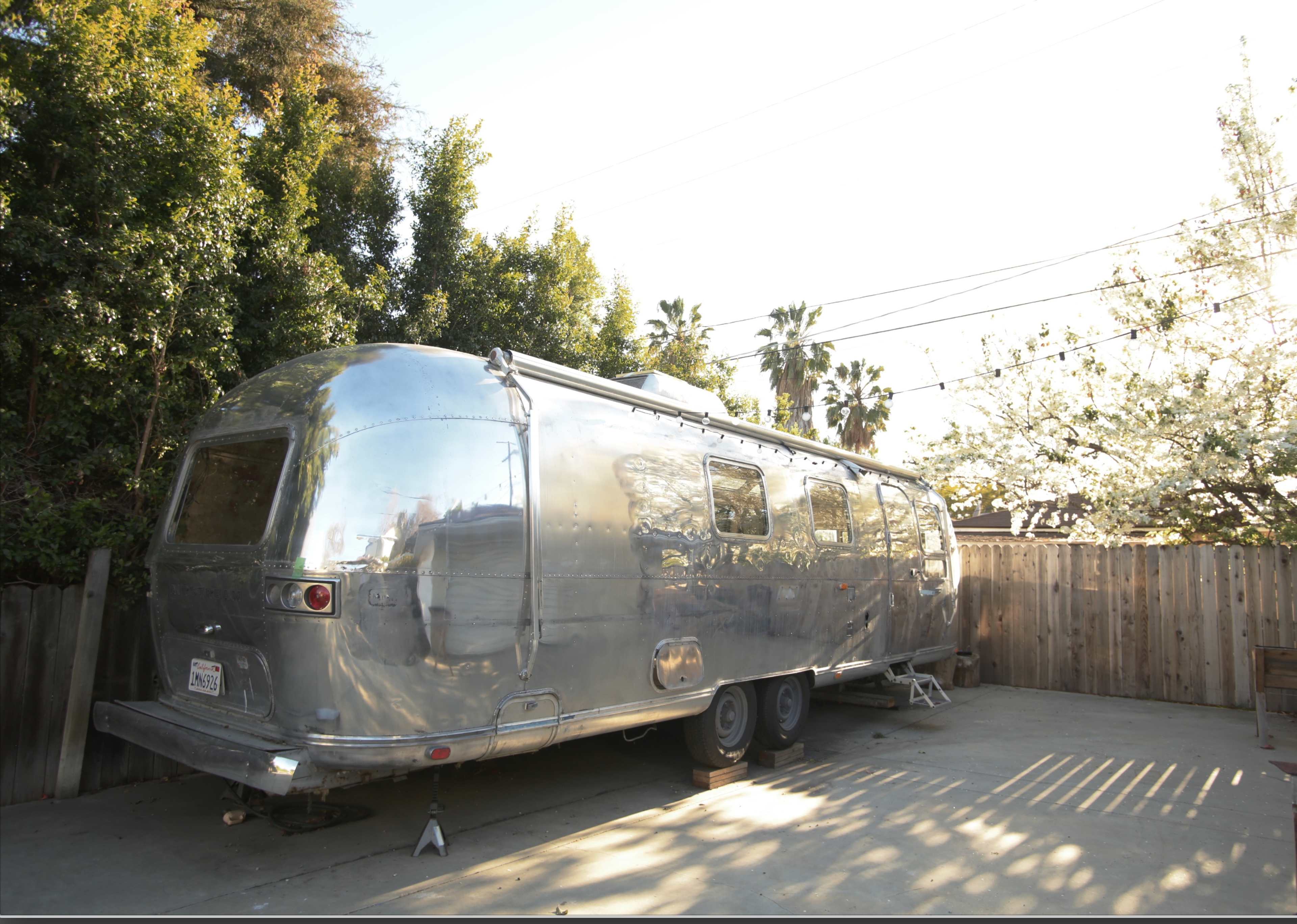 A shiny silver Airstream trailer is parked on a concrete pad surrounded by trees and a wooden fence.