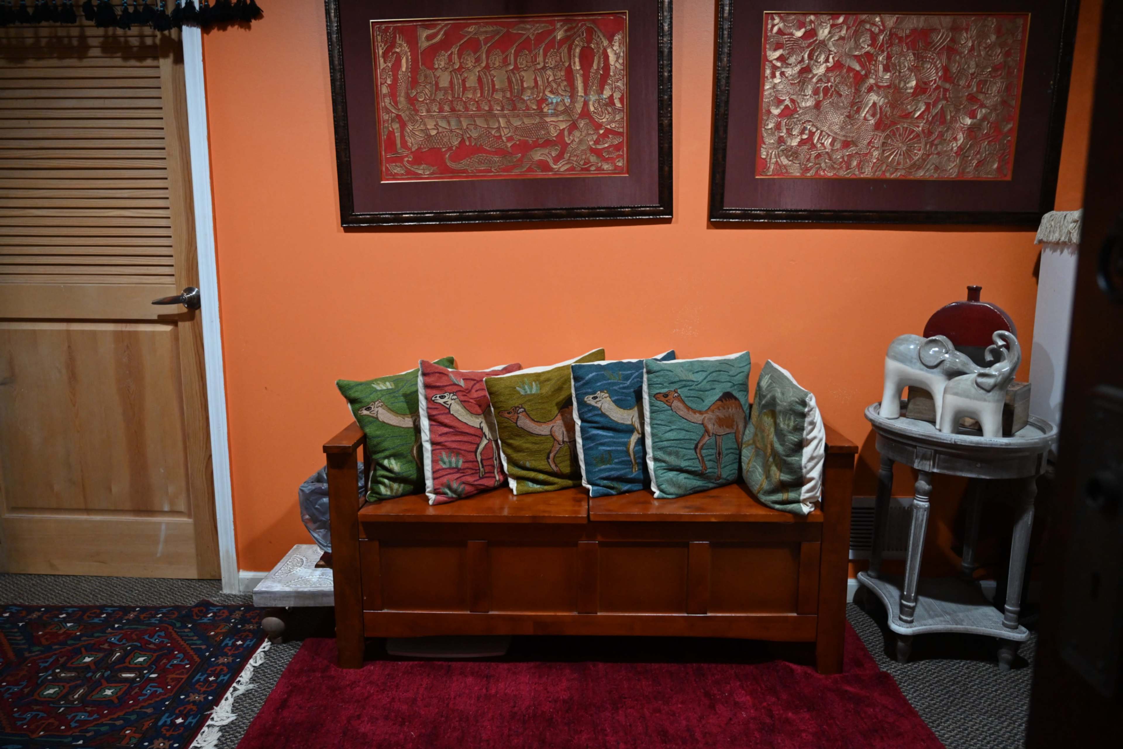 A wooden bench with colorful pillows featuring camel designs is positioned against an orange wall, adorned with framed artwork above it.