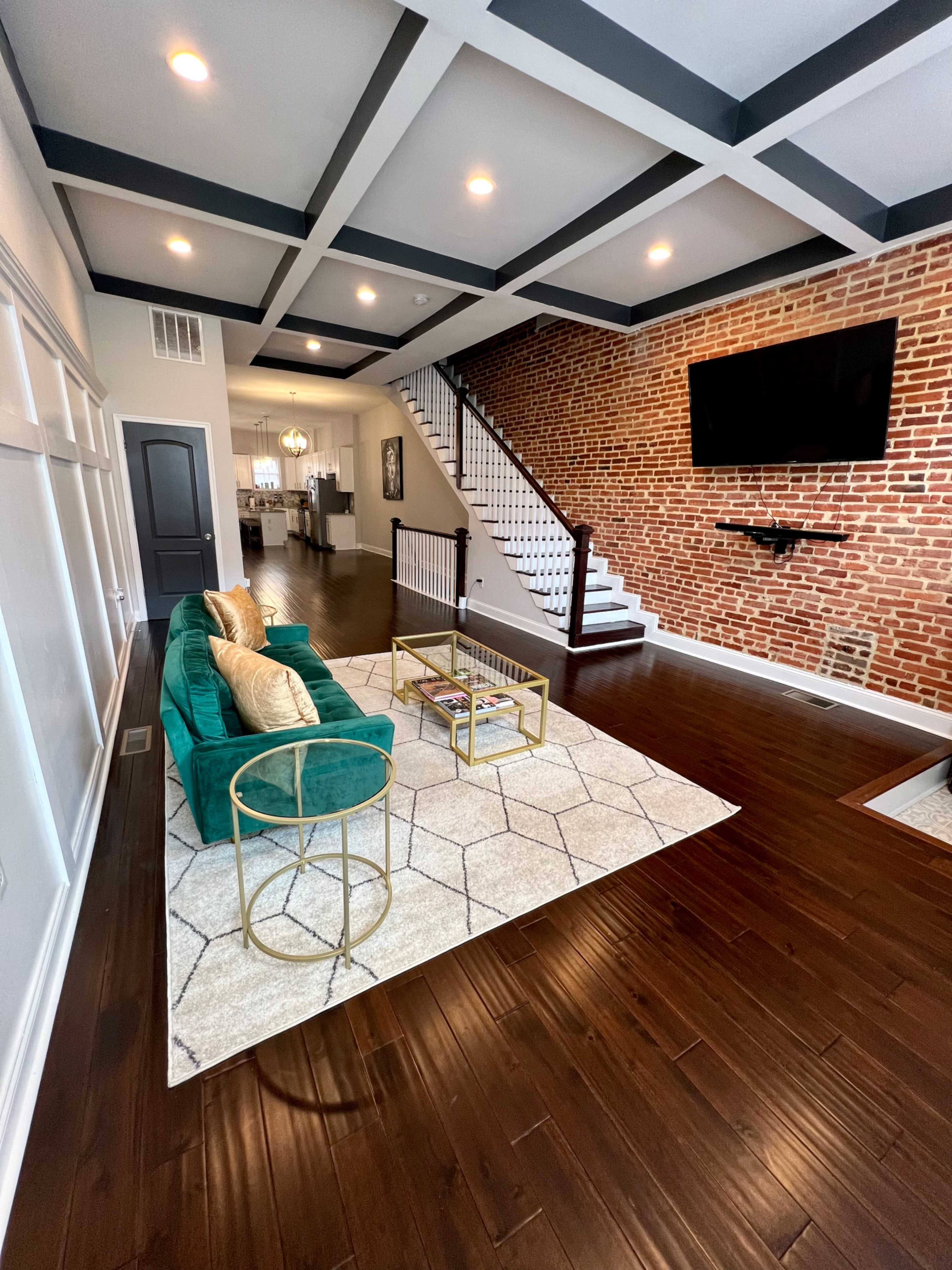 A modern living room featuring a green couch, a gold coffee table on a patterned rug, and a staircase leading to a second floor with exposed brick walls.
