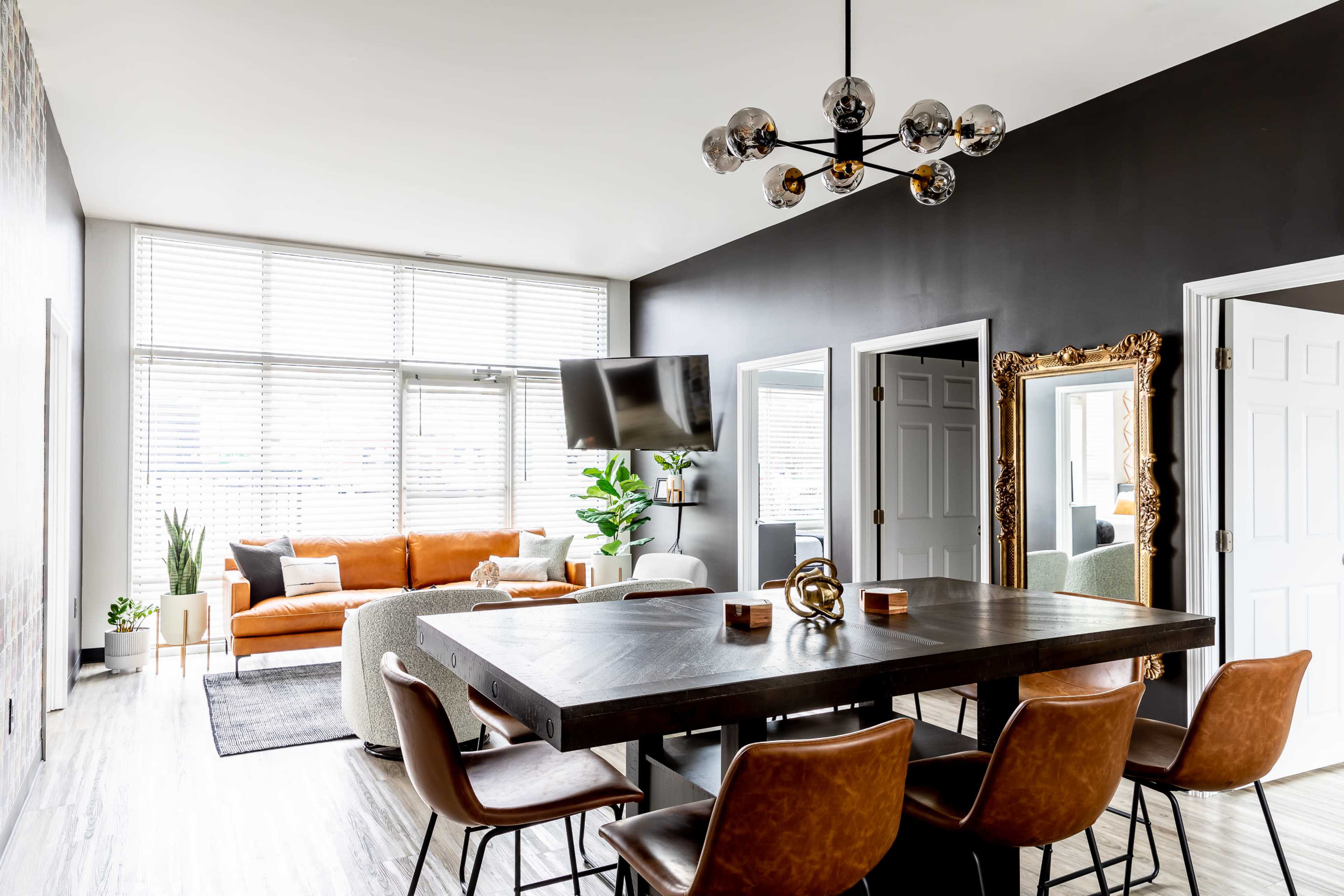 A modern living space features a dining area with a large table and chairs, a cozy orange sofa, a TV mounted on the wall, and large windows providing natural light.