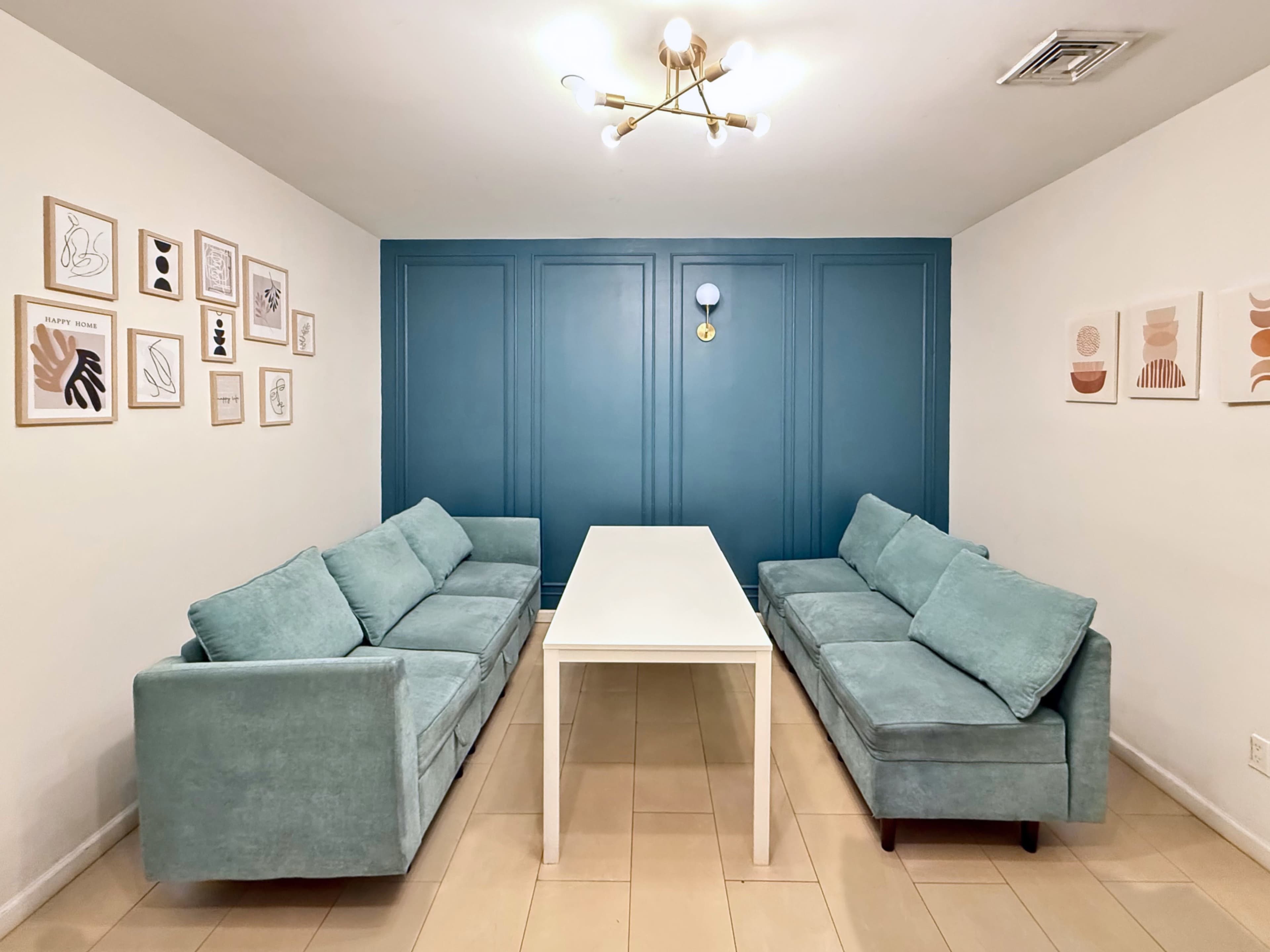 The scene features a modern meeting room with two light blue couches flanking a white rectangular table against a wall painted in a deep teal color, adorned with framed artwork.