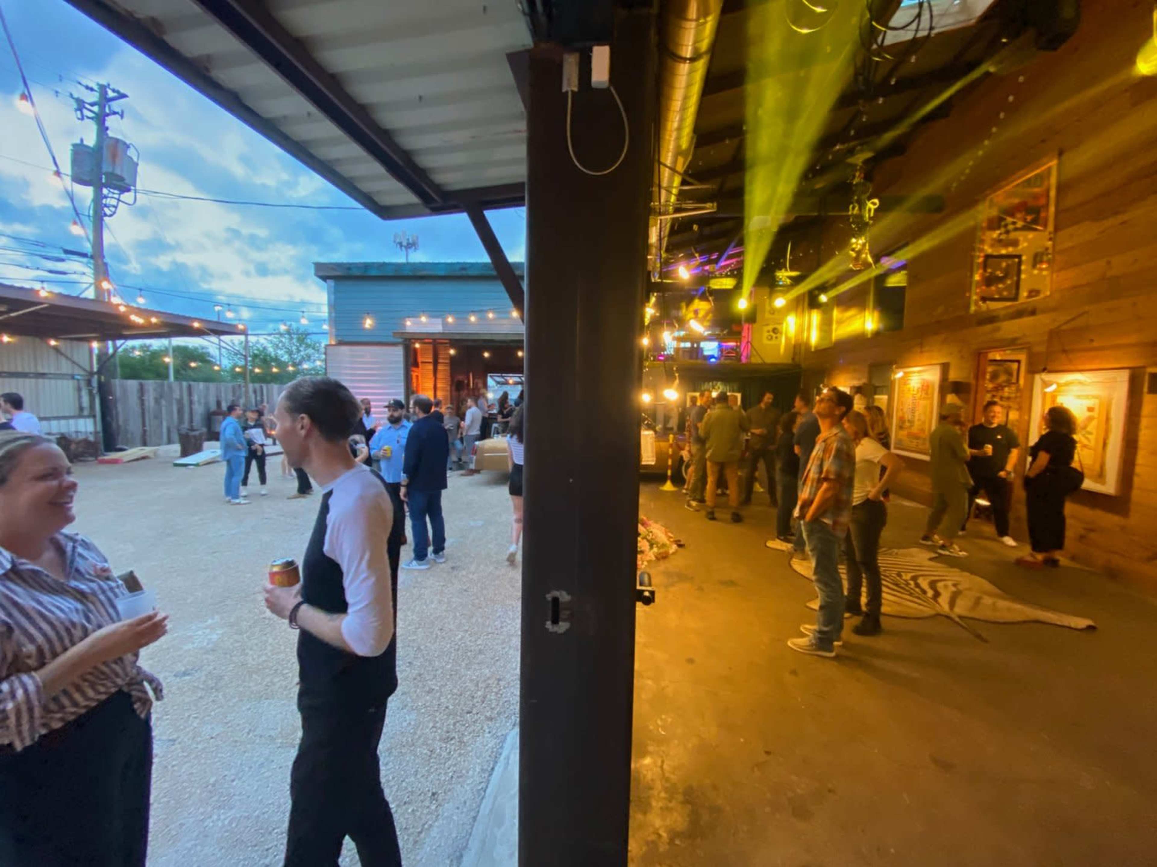 A group of people socializes at an outdoor event space with string lights and a rustic atmosphere.