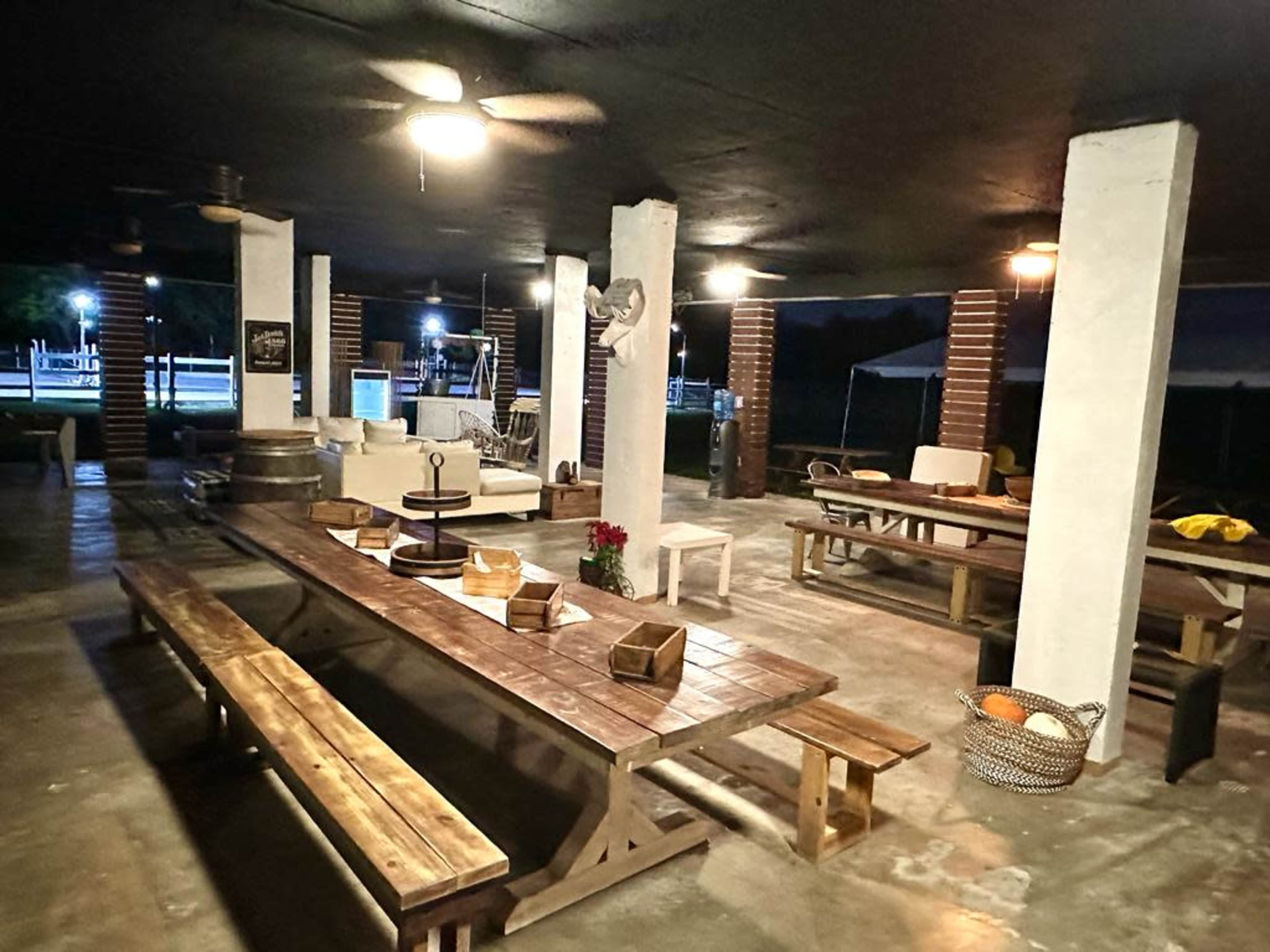 The image shows a spacious outdoor gathering area with multiple wooden tables, benches, and a few lounge chairs under a covered roof, illuminated by ceiling fans and ambient lighting.