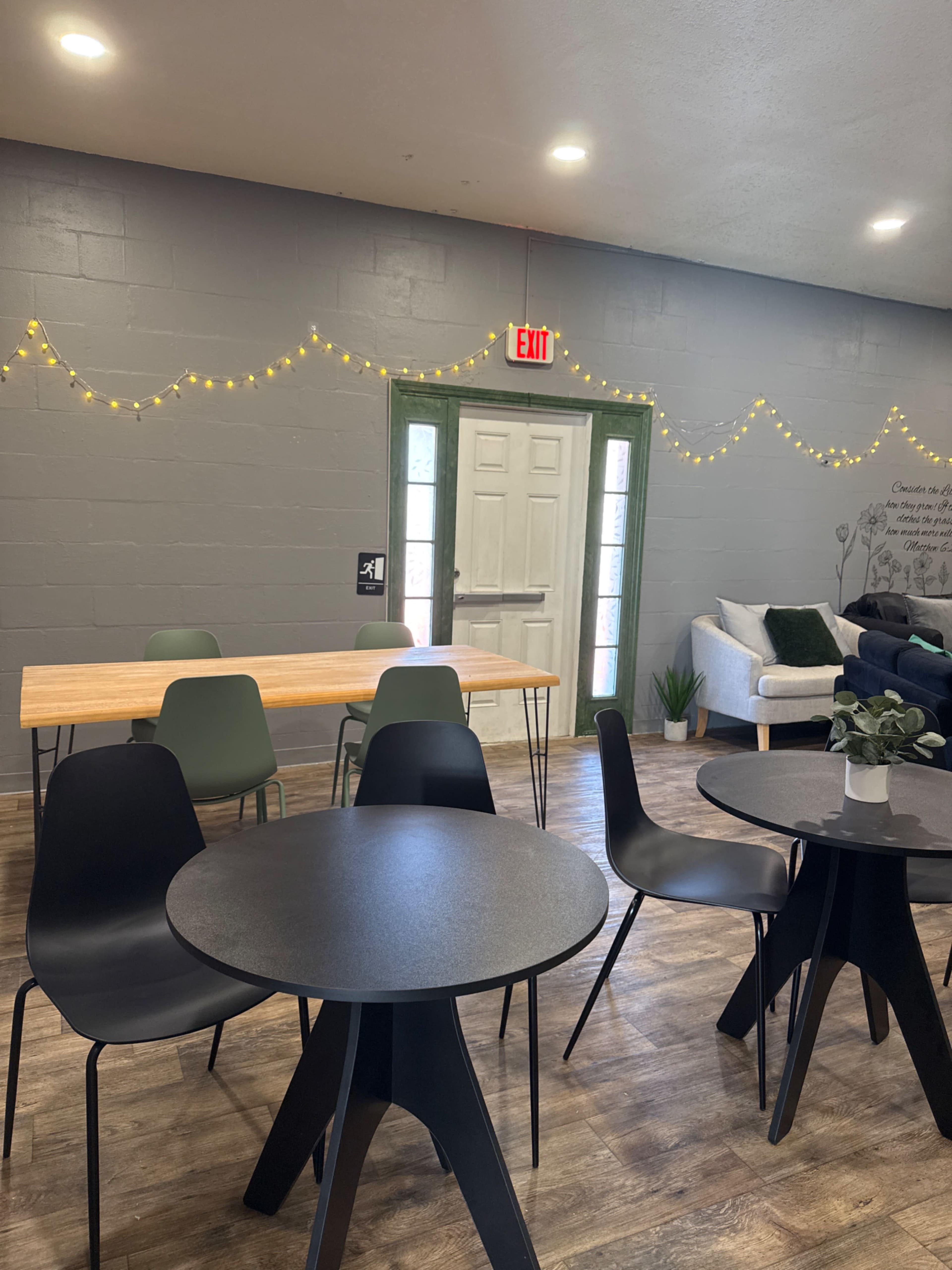 The image shows a cozy room featuring several black and green chairs around black and wooden tables, with string lights adorning the walls and a door leading outside.