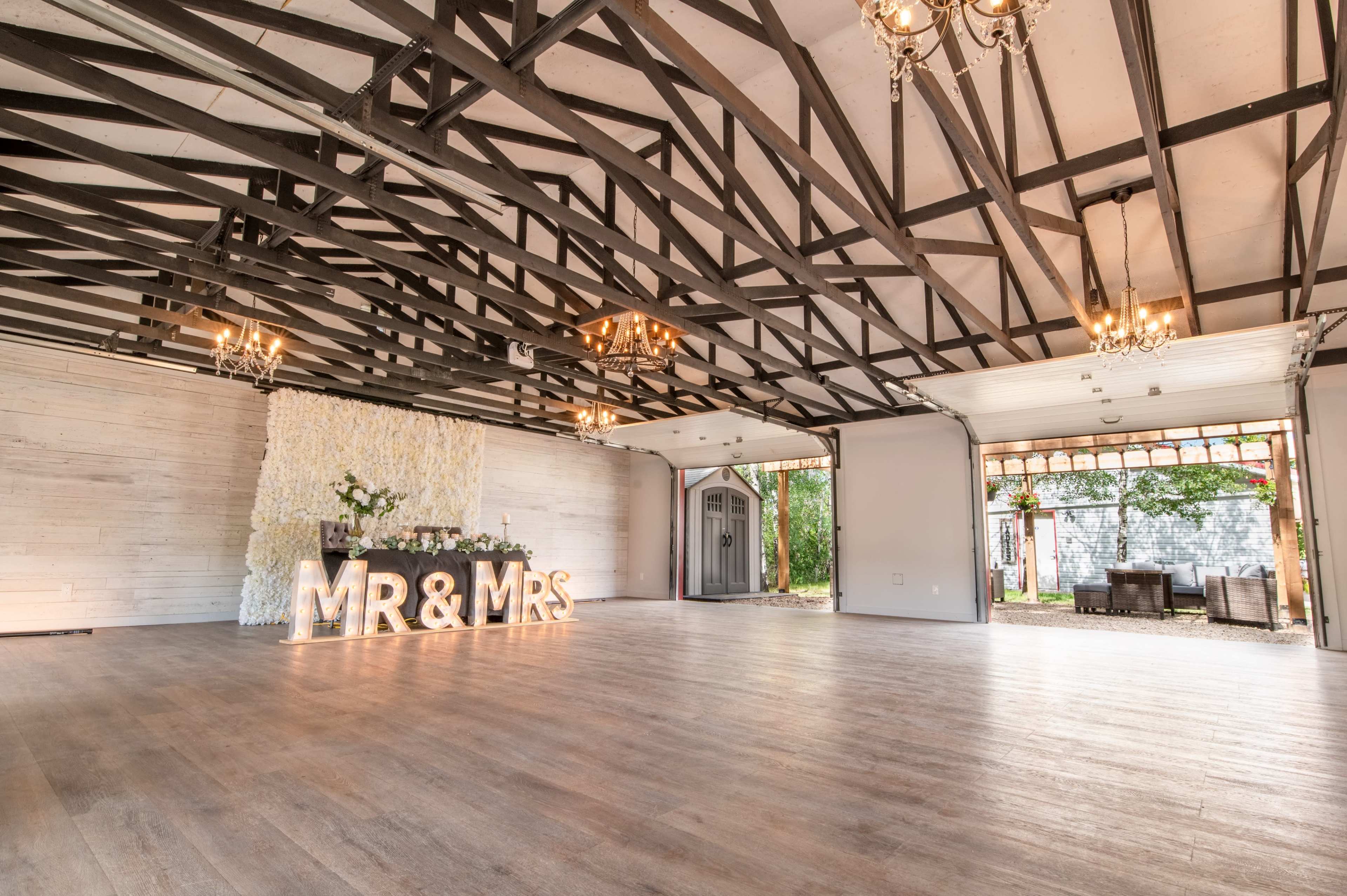 The image shows a spacious event venue featuring a decorative backdrop with the text "MR & MRS," elegant chandeliers, and wooden beams in the ceiling.