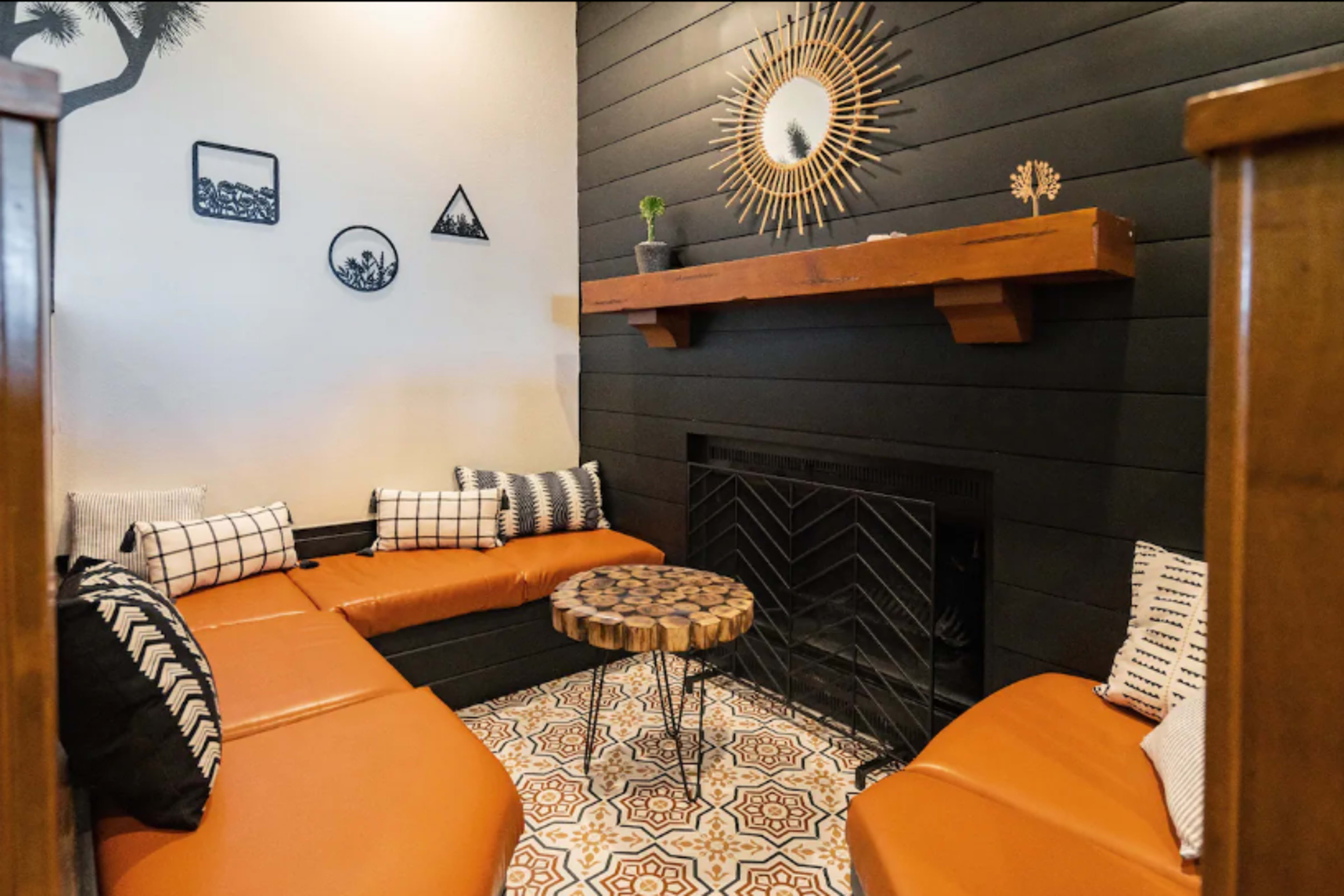 The image shows a cozy seating area with an orange sectional couch, a round wooden coffee table, and a black fireplace, decorated with framed art and a mirror on the wall.
