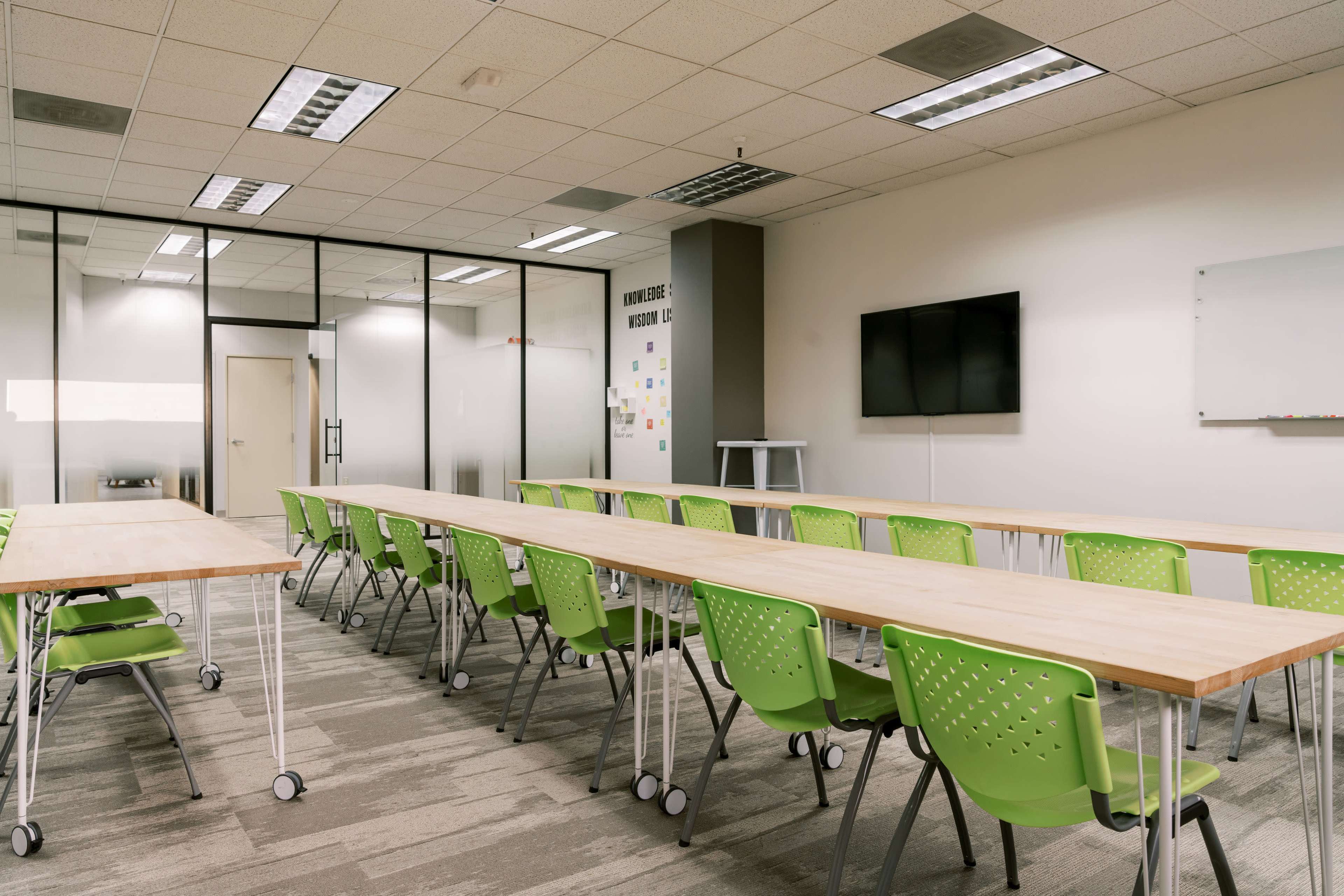A conference room with long tables arranged in a U-shape and green chairs, featuring a television and whiteboard on the walls.