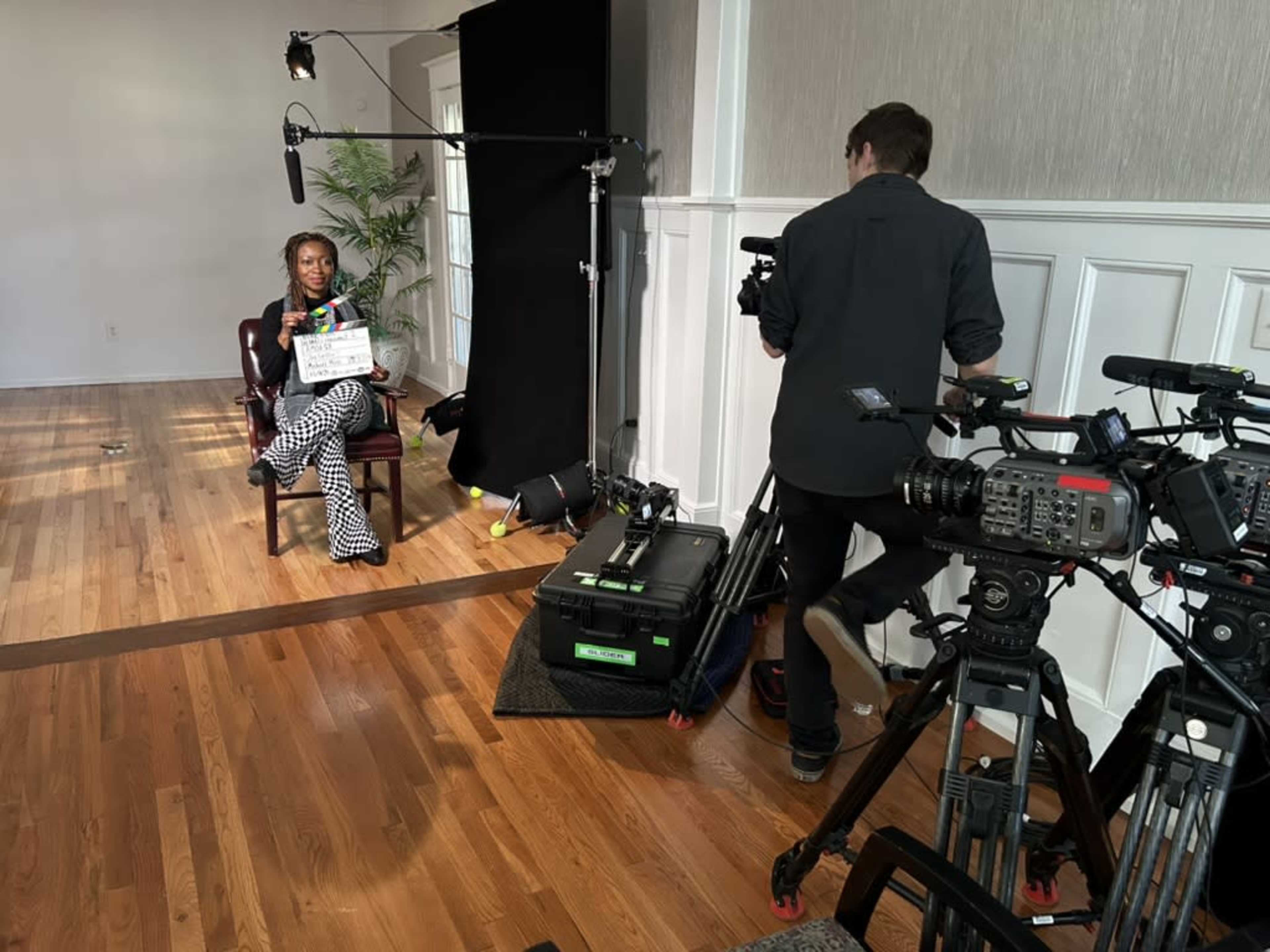 A woman sits in a chair holding a clapperboard while a man operates a camera nearby in a film set environment.