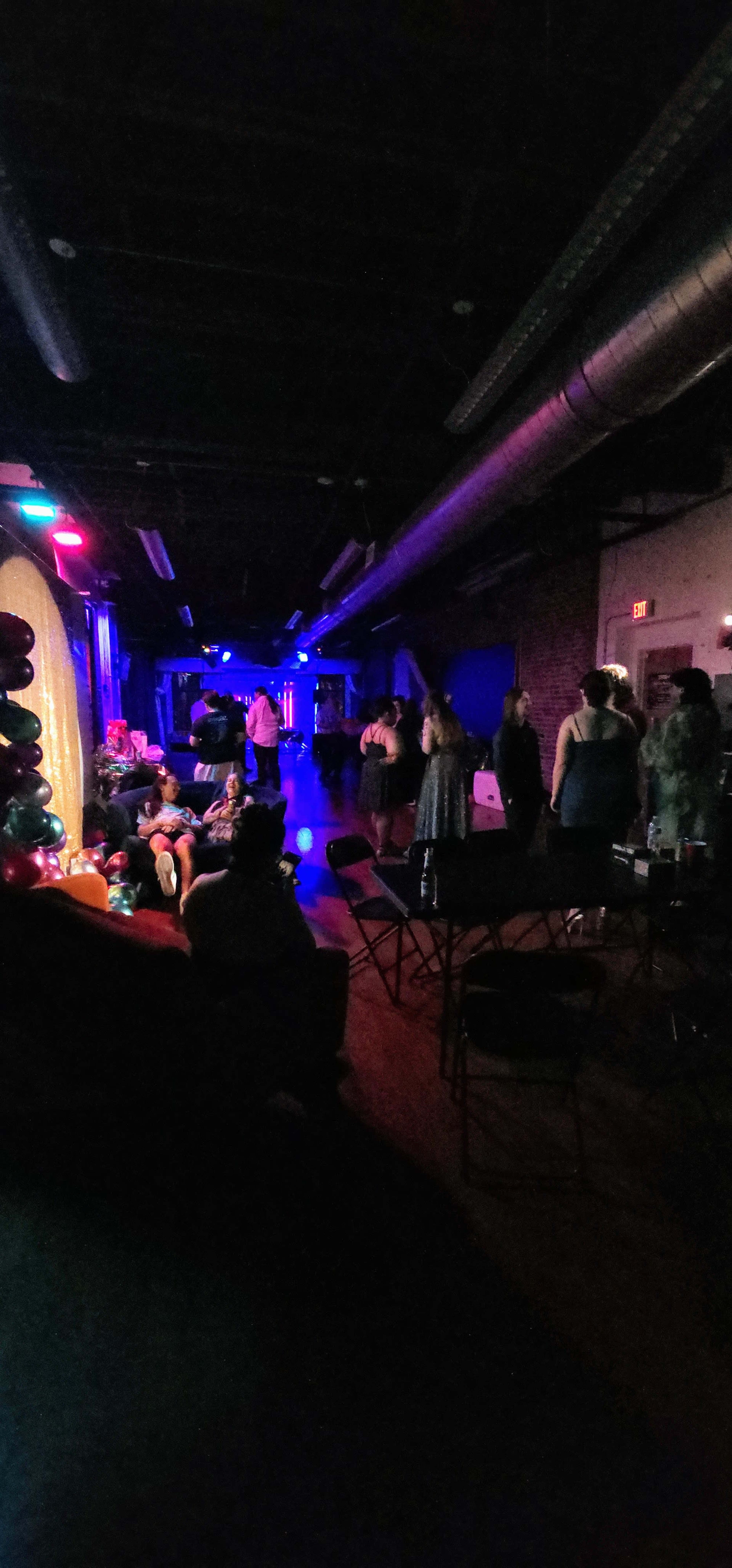 The image shows a dimly lit venue with colorful lights, where people are socializing and dancing.