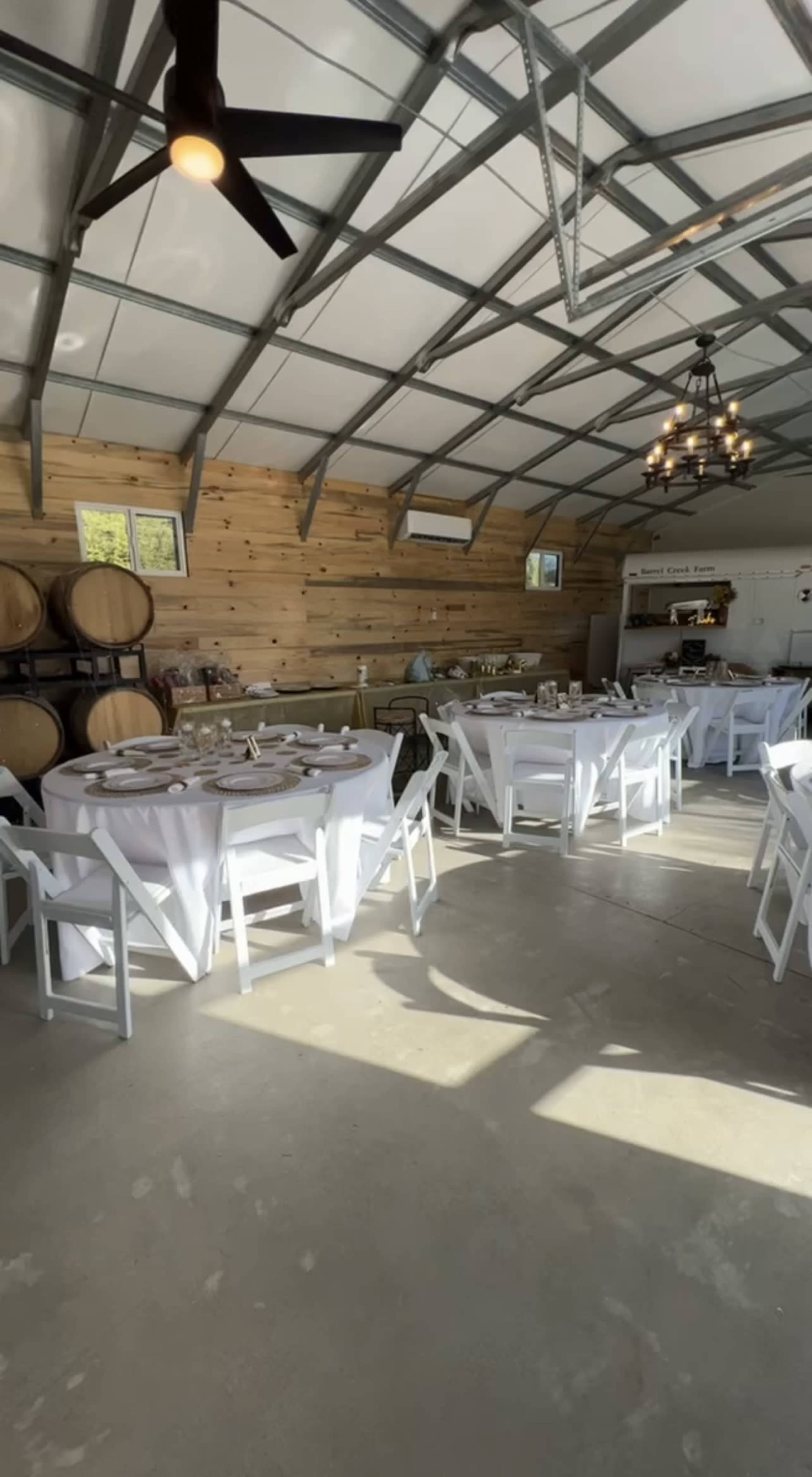 A rustic dining space with multiple round tables set with white tablecloths, surrounded by wooden walls and wine barrels.