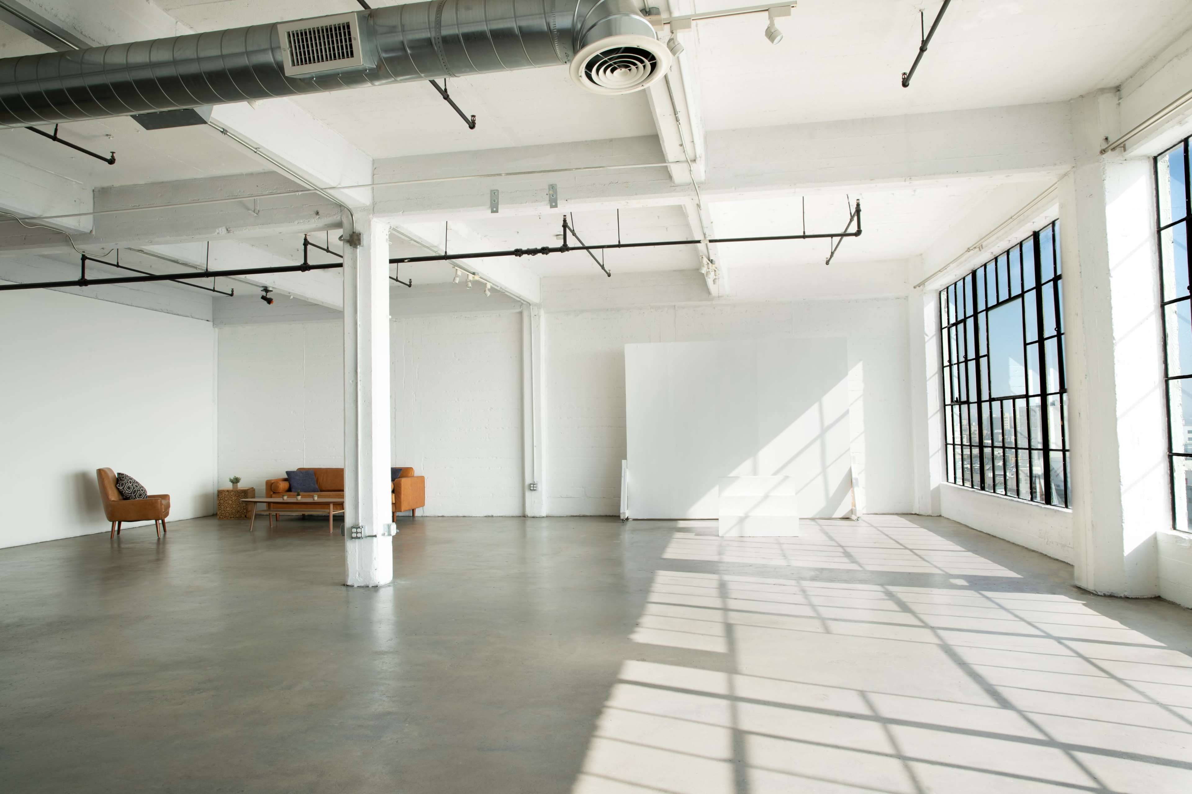 The image shows a spacious, empty industrial-style room with large windows, concrete flooring, and minimal furnishings, including an orange couch and a chair.