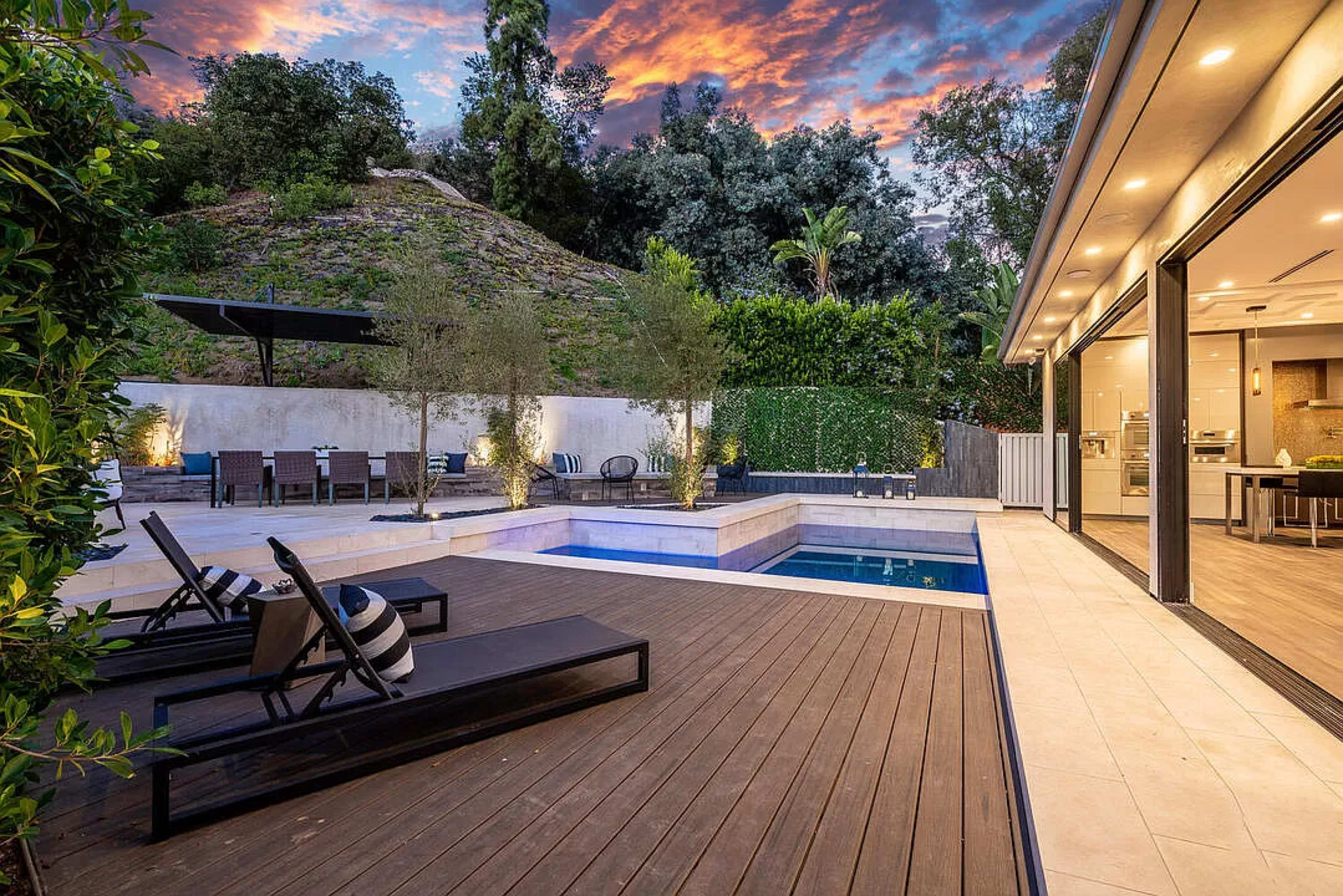 The image shows a modern backyard featuring a pool, wooden deck, lounge chairs, and a landscaped hillside backdrop under an evening sky.