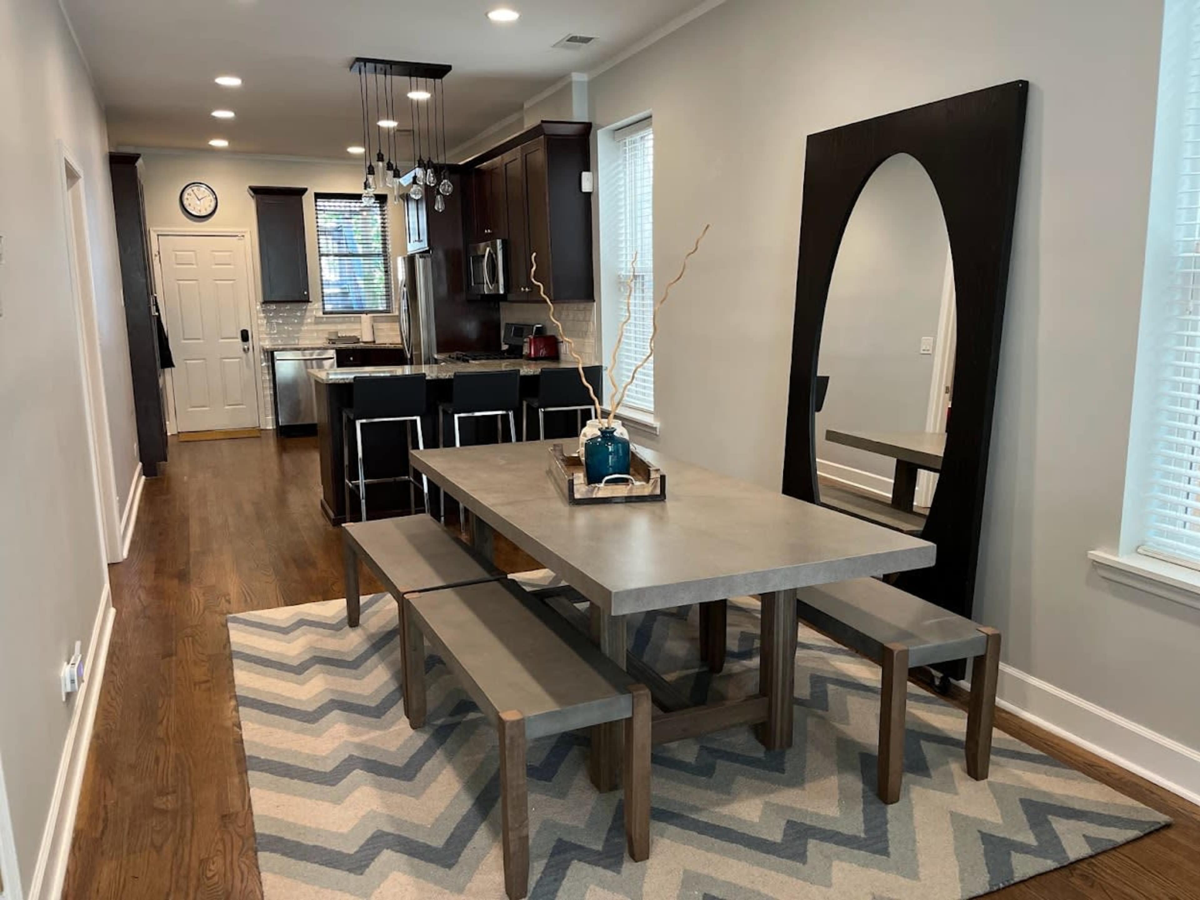 A modern dining area features a large concrete table with wooden benches, surrounded by a rug with a chevron pattern, alongside a kitchen space with dark cabinets.