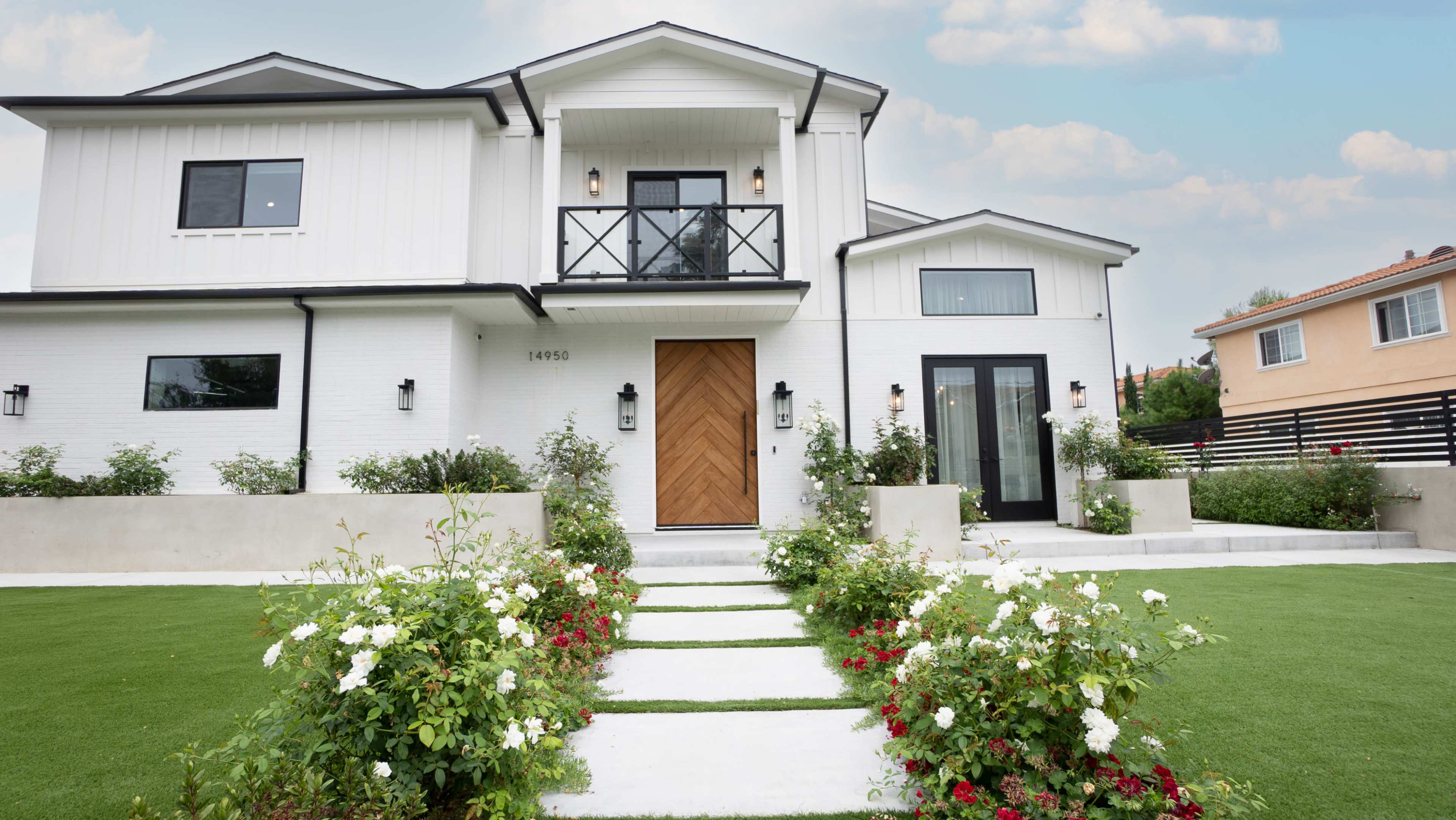 A modern, two-story white house with a large wooden front door, surrounded by neatly arranged flower beds and a well-maintained lawn.