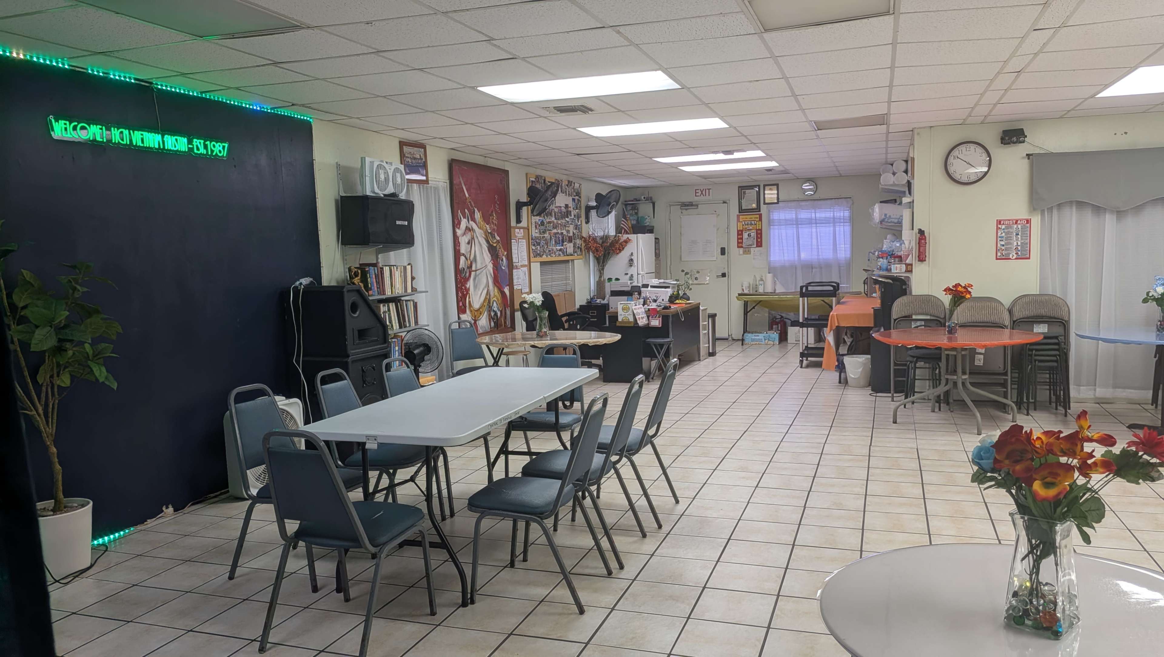 The image shows a spacious interior of a community center with tables and chairs arranged for meetings, a reception area, and various decorations on the walls.