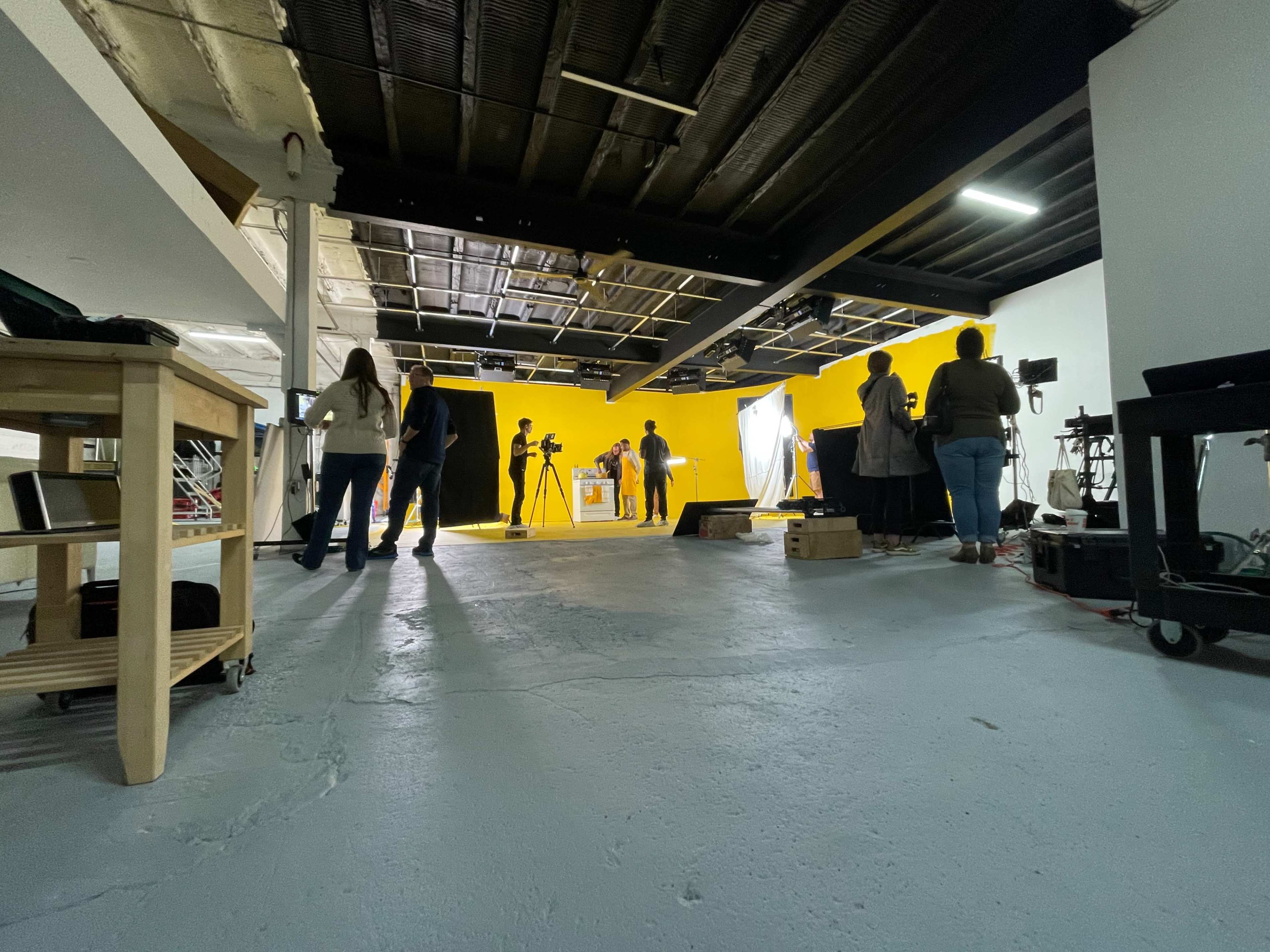 A photography studio features a bright yellow wall, with several people setting up equipment and preparing for a shoot.