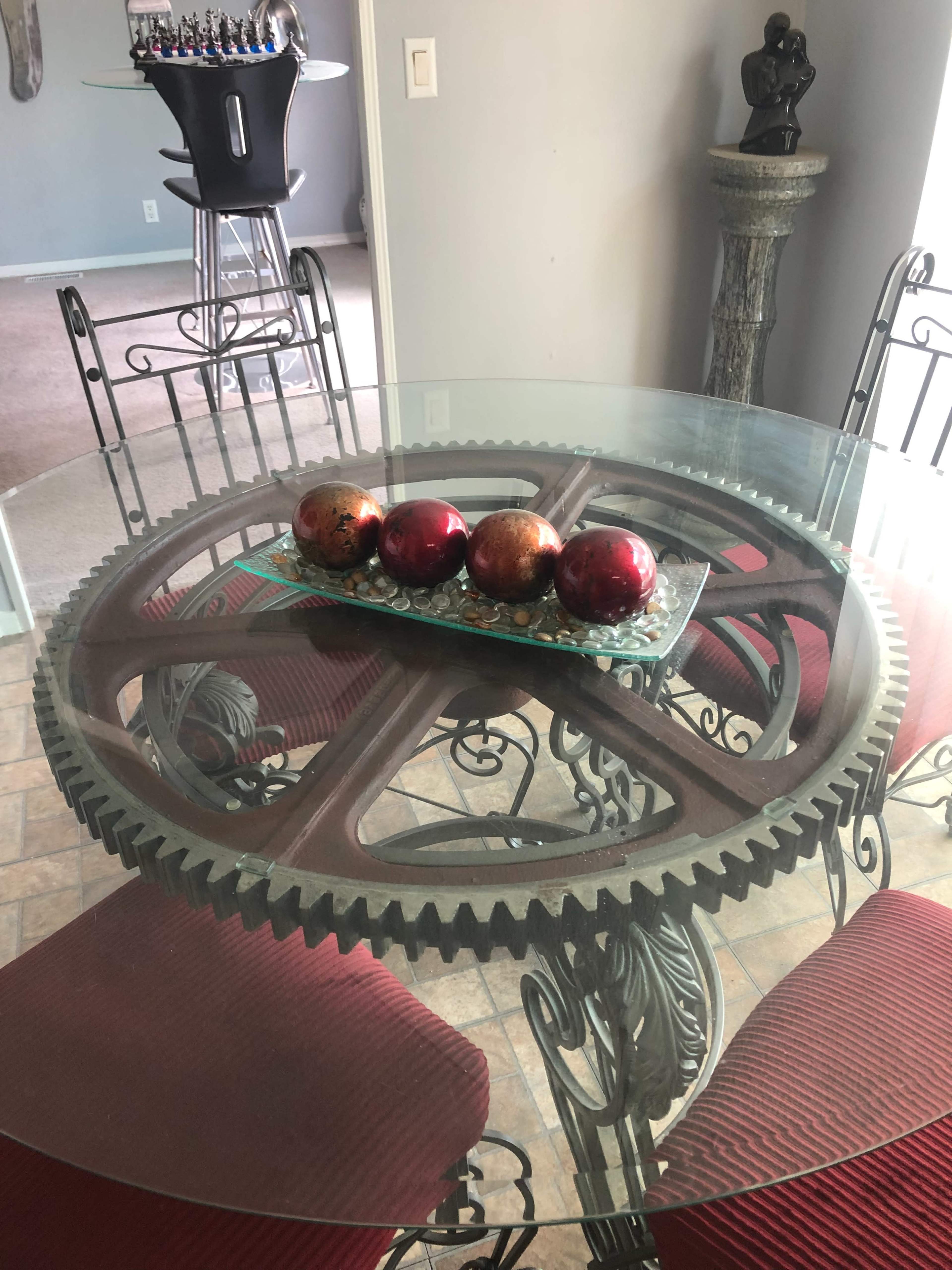 A glass-topped table with a gear-shaped base holds a decorative tray featuring four spherical ornaments, while red upholstered chairs surround it.