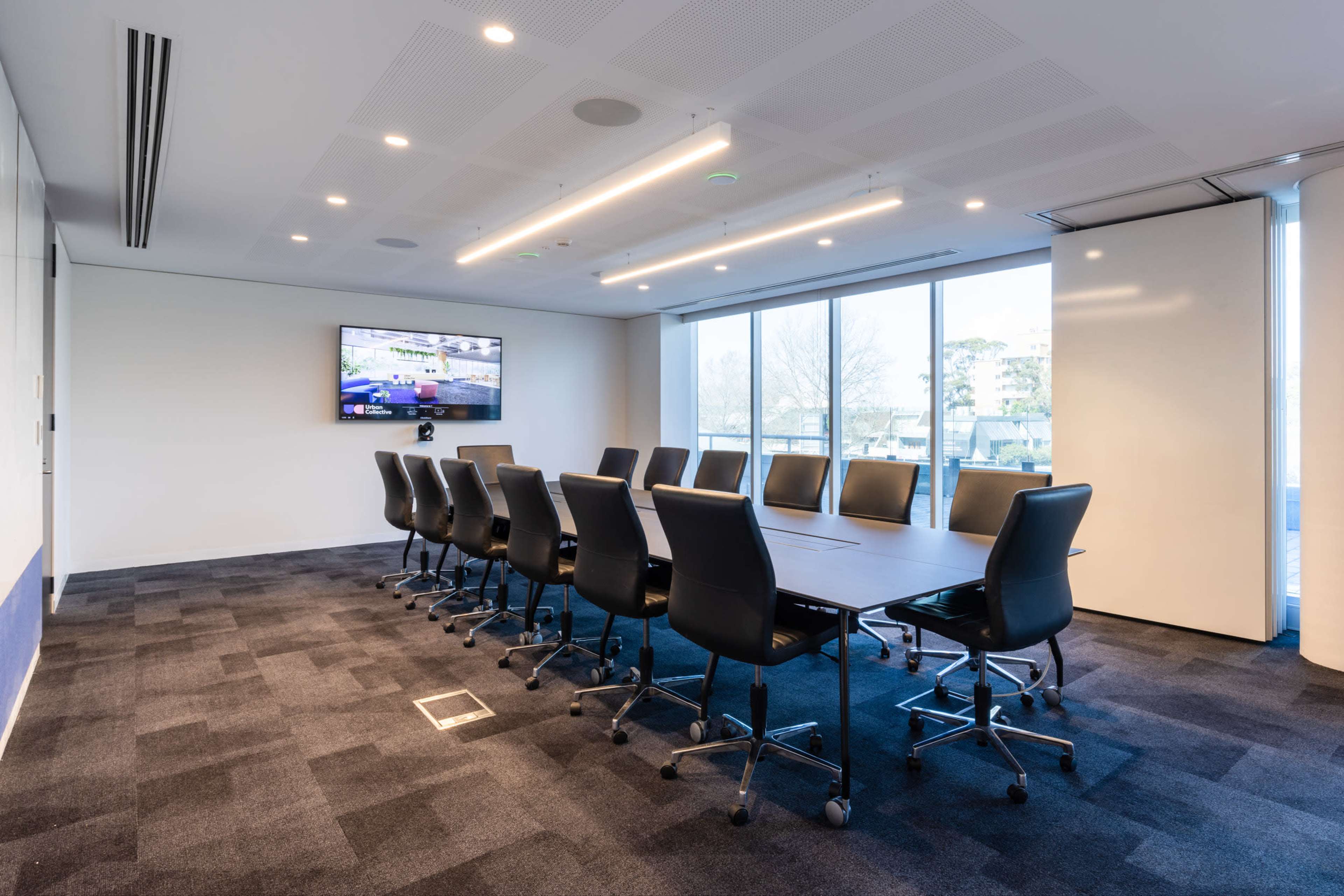 A modern conference room features a long table surrounded by black chairs, with a television mounted on the wall and large windows allowing natural light.