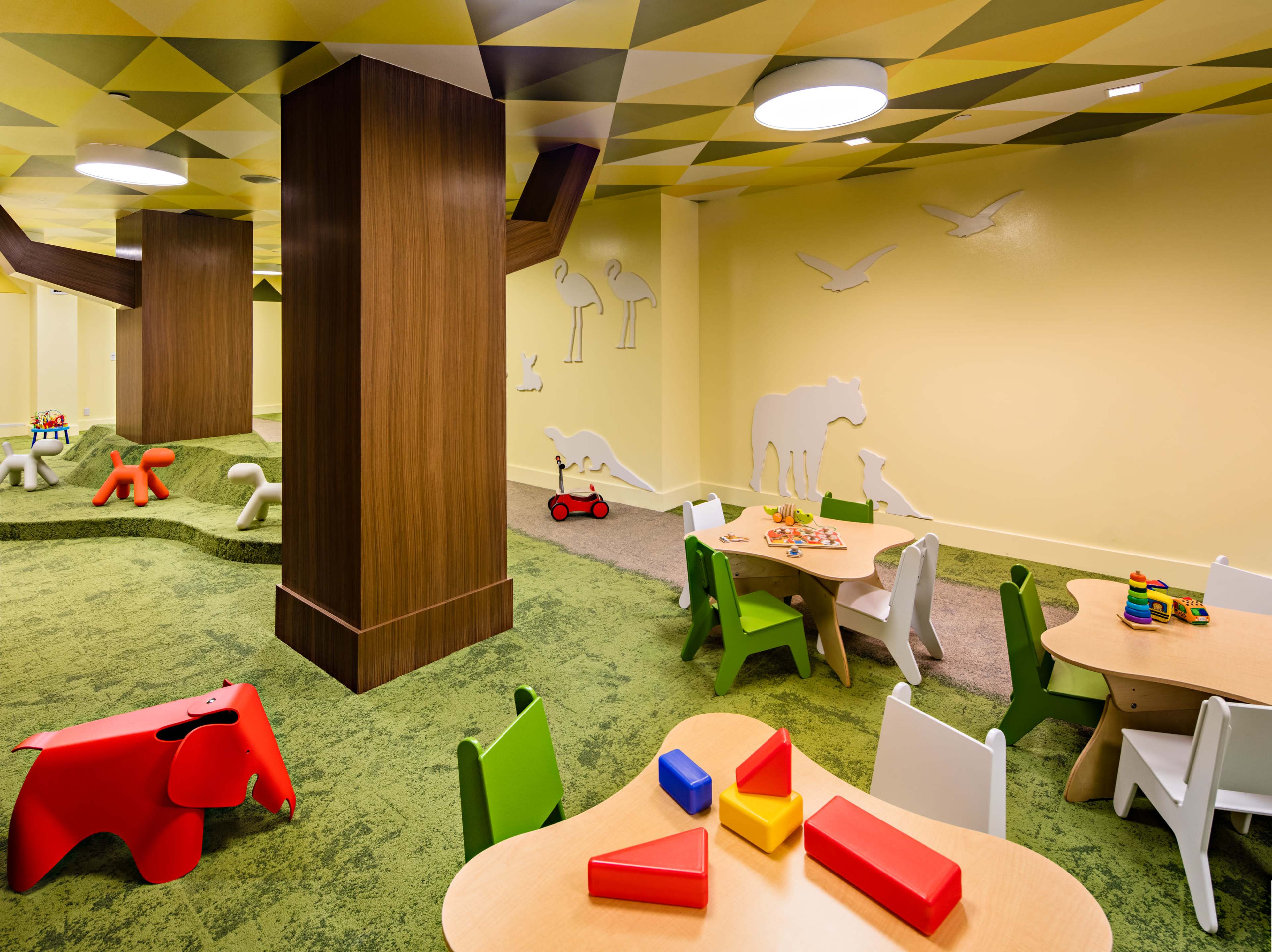 The image shows a brightly colored children's playroom with green carpet, animal-shaped play furniture, and tables with toys.