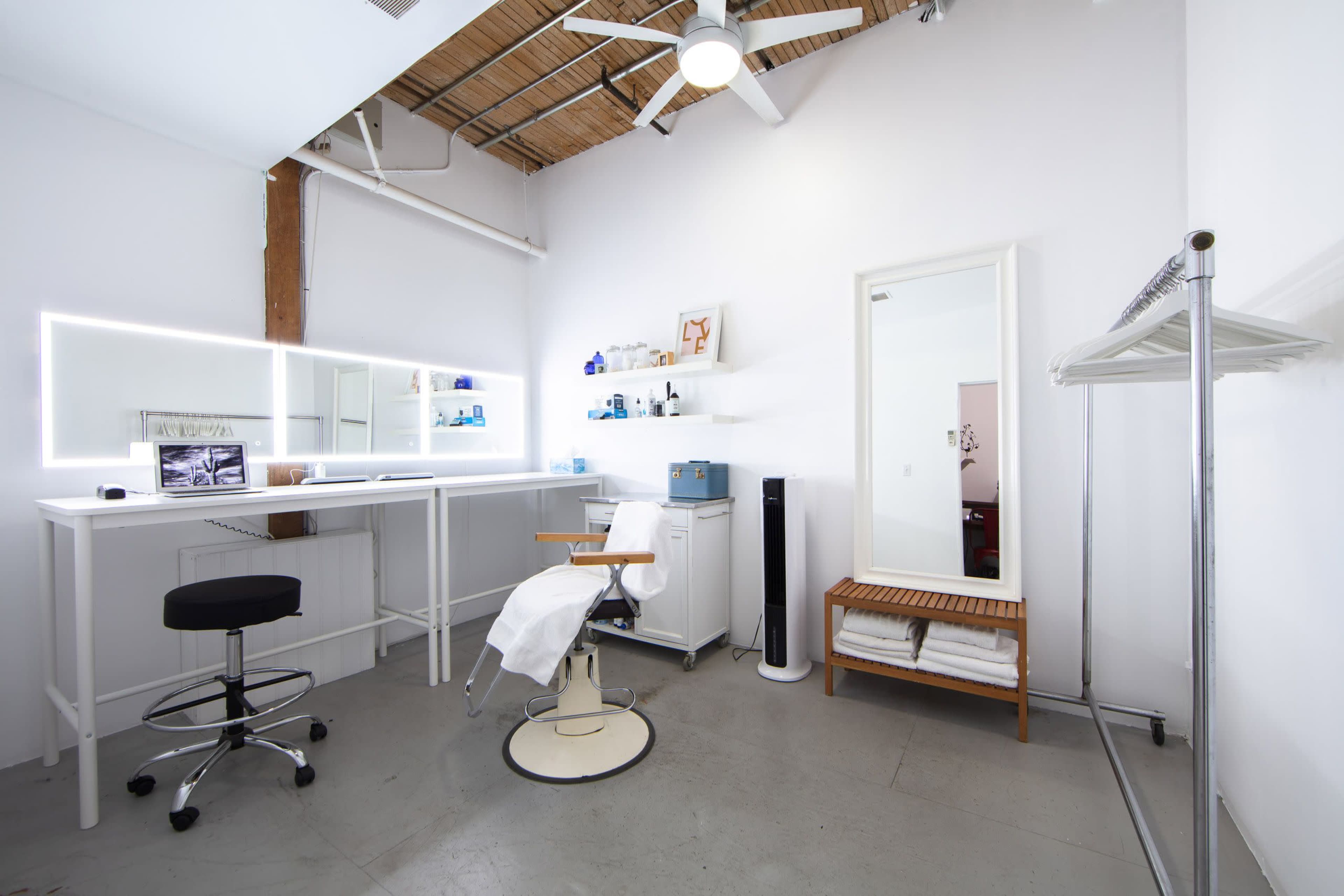A minimalist beauty studio featuring a styling chair, a large mirror with backlighting, a storage shelf with various products, and a clothing rack.