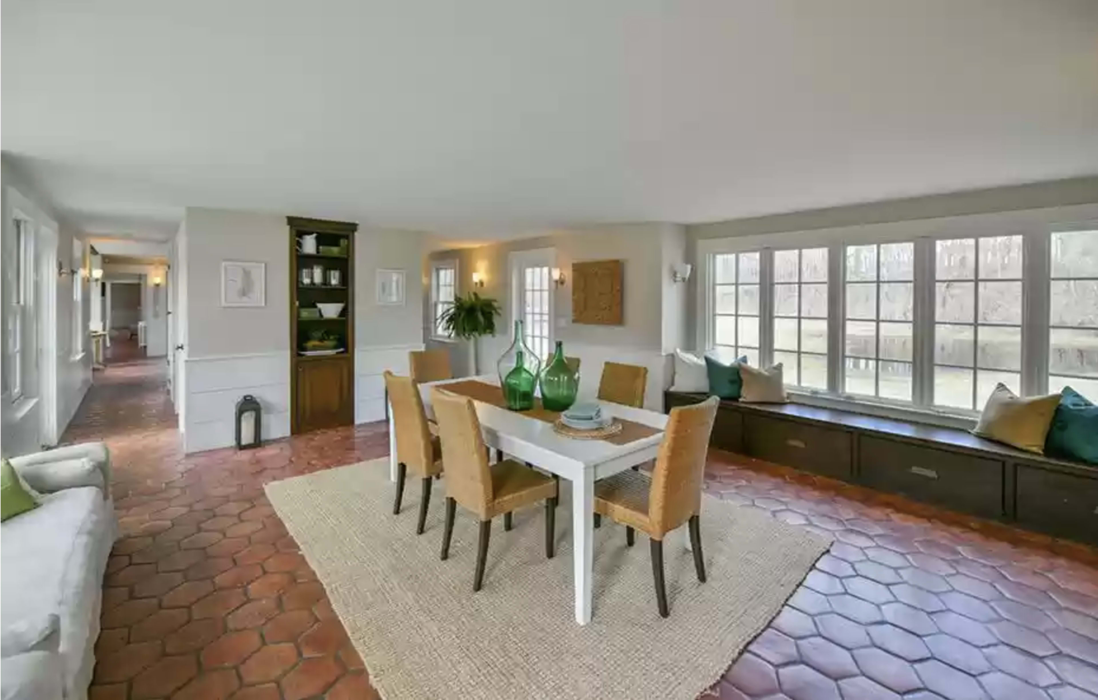 The image shows a well-lit dining area with a white table surrounded by four chairs, with a built-in bench and large windows letting in natural light.