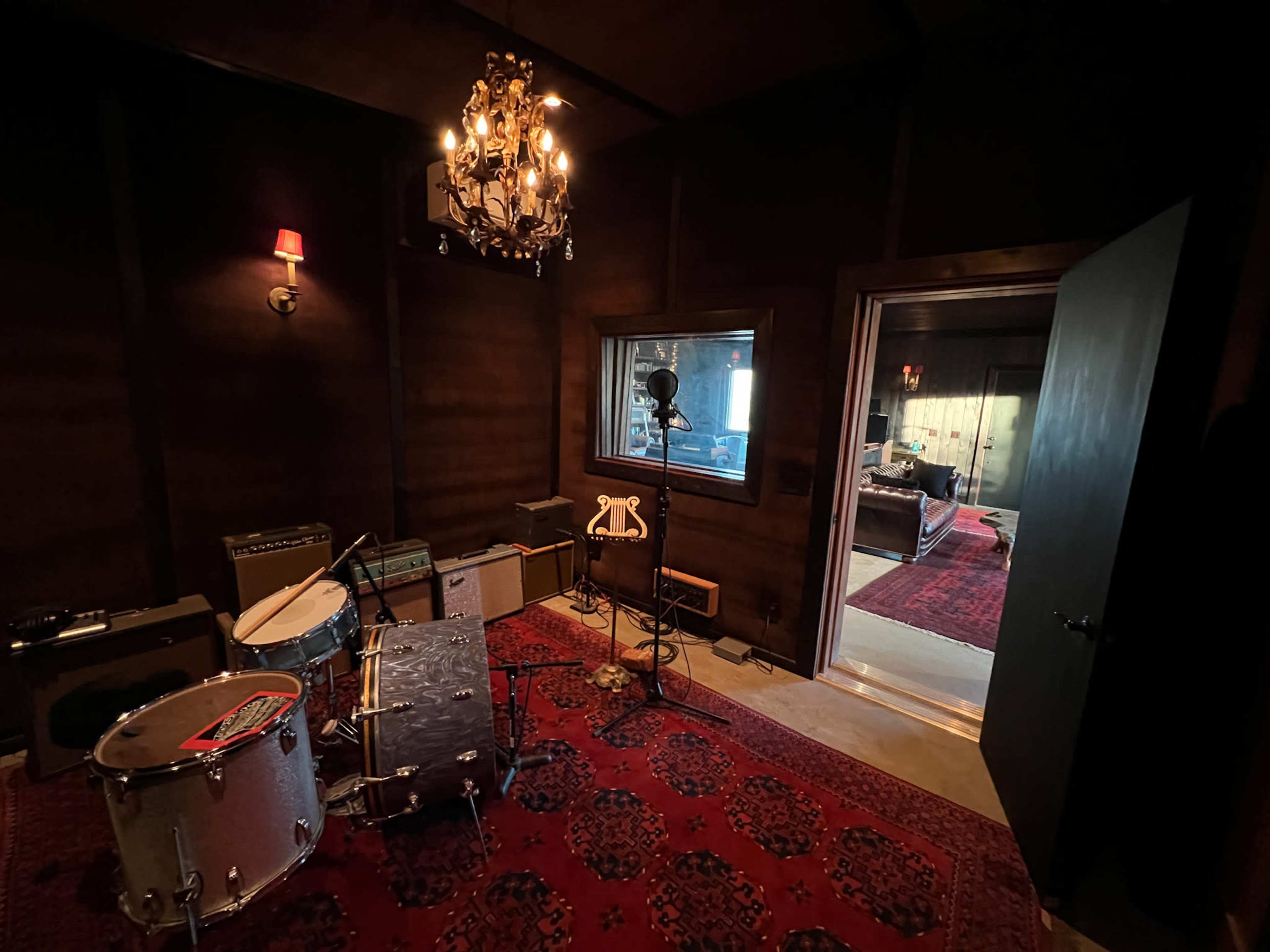 The image shows a dimly lit recording studio room with a drum set, amplifiers, and a microphone stand, leading to an adjacent room visible through an open door.