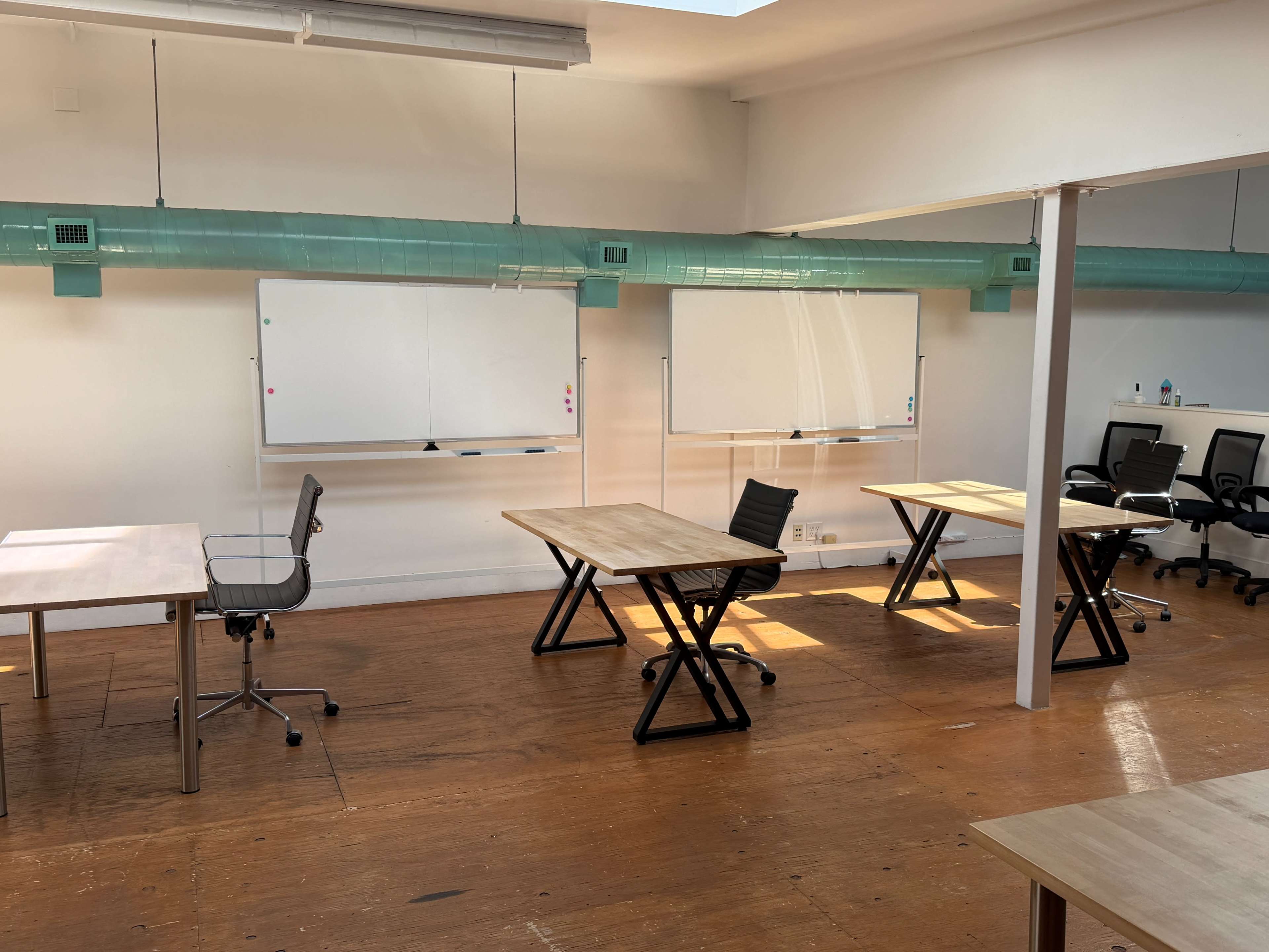 The image shows an empty office space with several wooden tables and chairs, along with whiteboards on the walls.