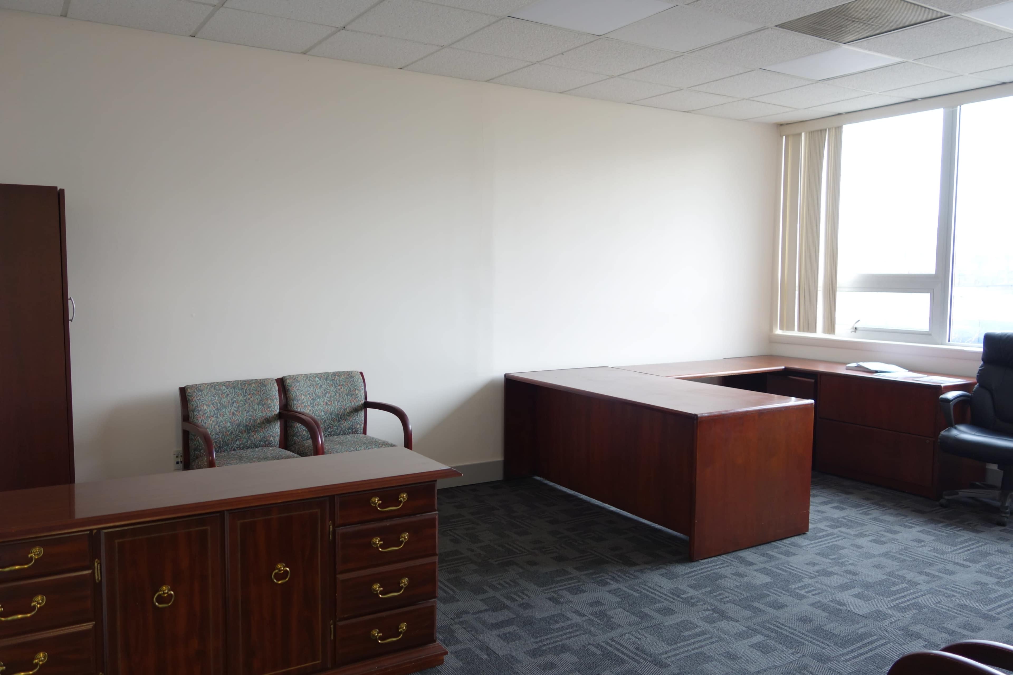 A sparsely furnished office with two wooden desks, a filing cabinet, and two chairs along one wall.