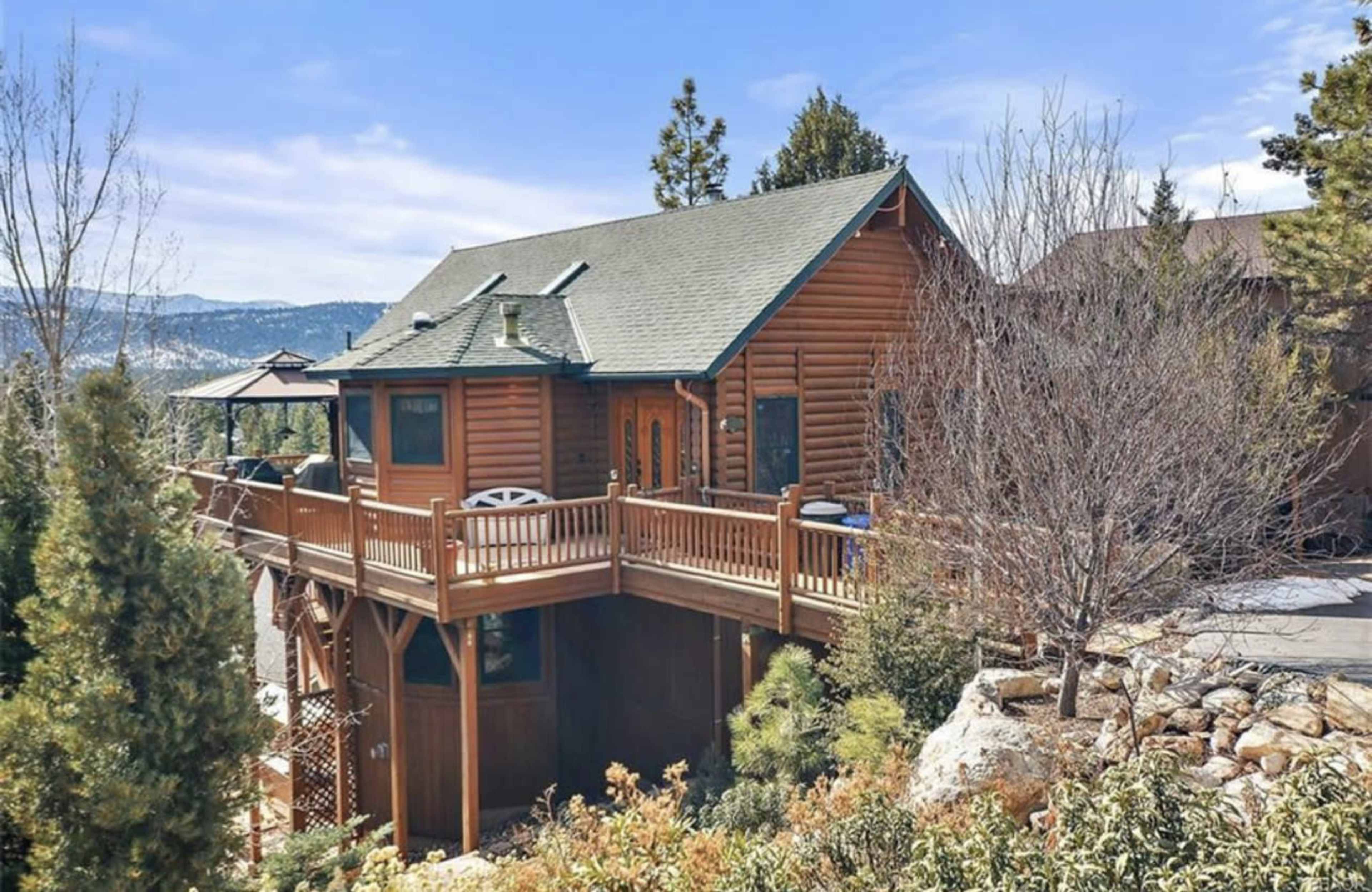 A wooden cabin with a green roof sits elevated on a hillside, surrounded by trees and mountains in the background.