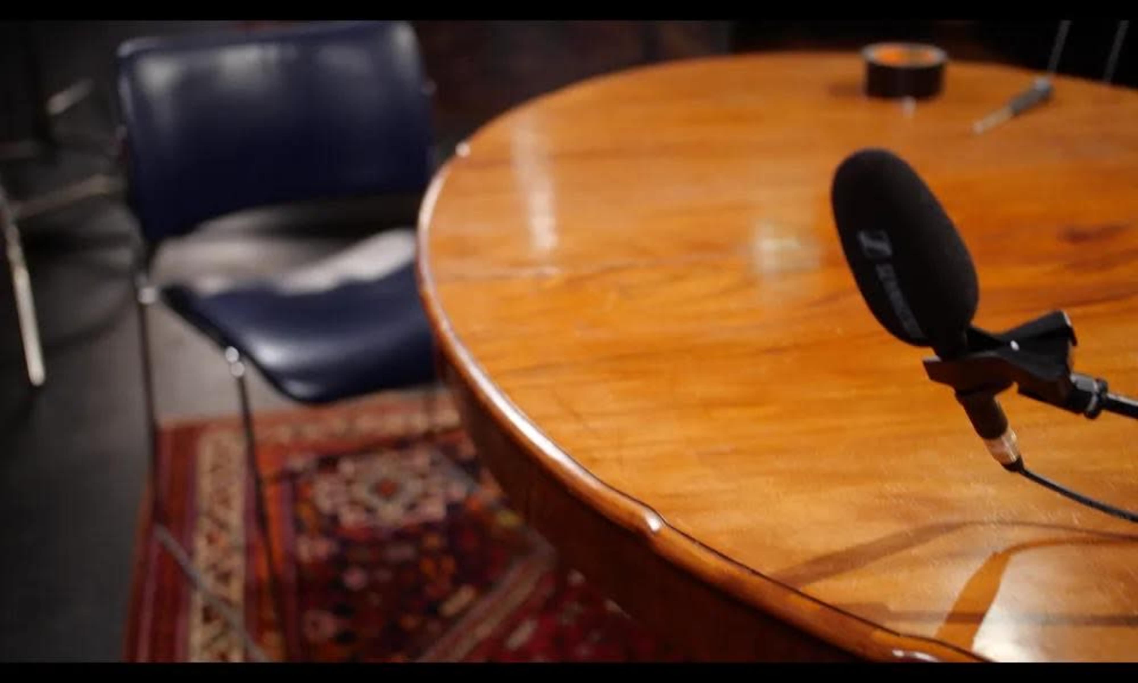 The image shows a wooden table with a microphone positioned on it, accompanied by a blue chair and a patterned rug beneath.