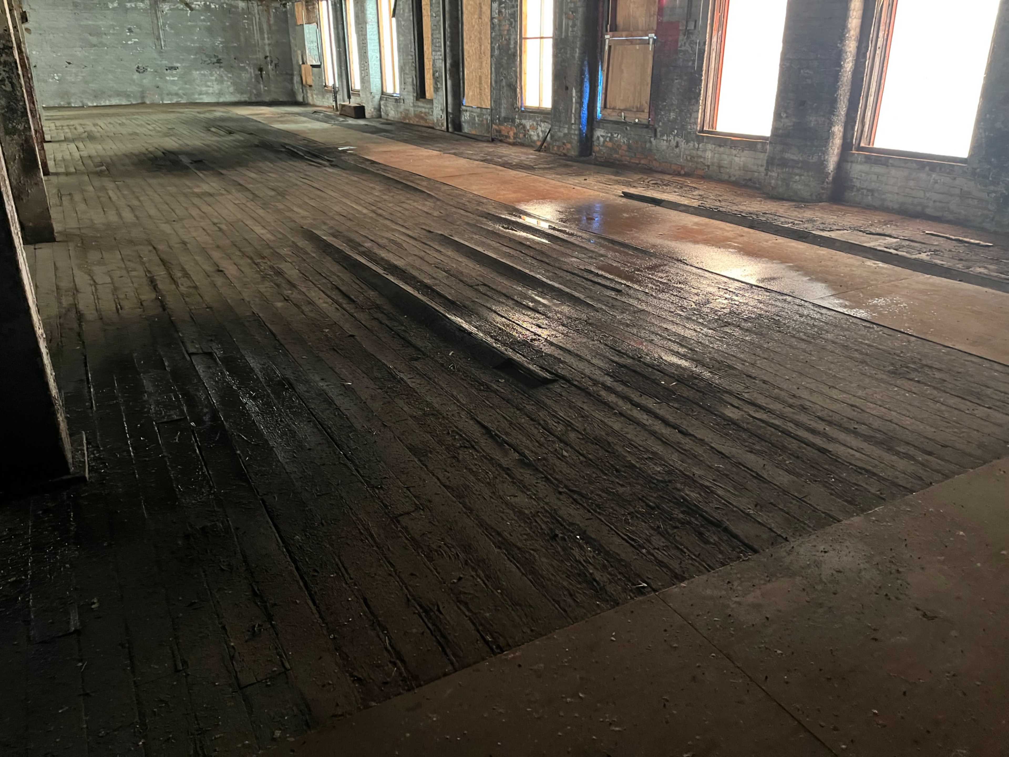 The image shows a dimly lit, empty interior space with wooden flooring that appears worn and dirty, featuring large windows along the walls.
