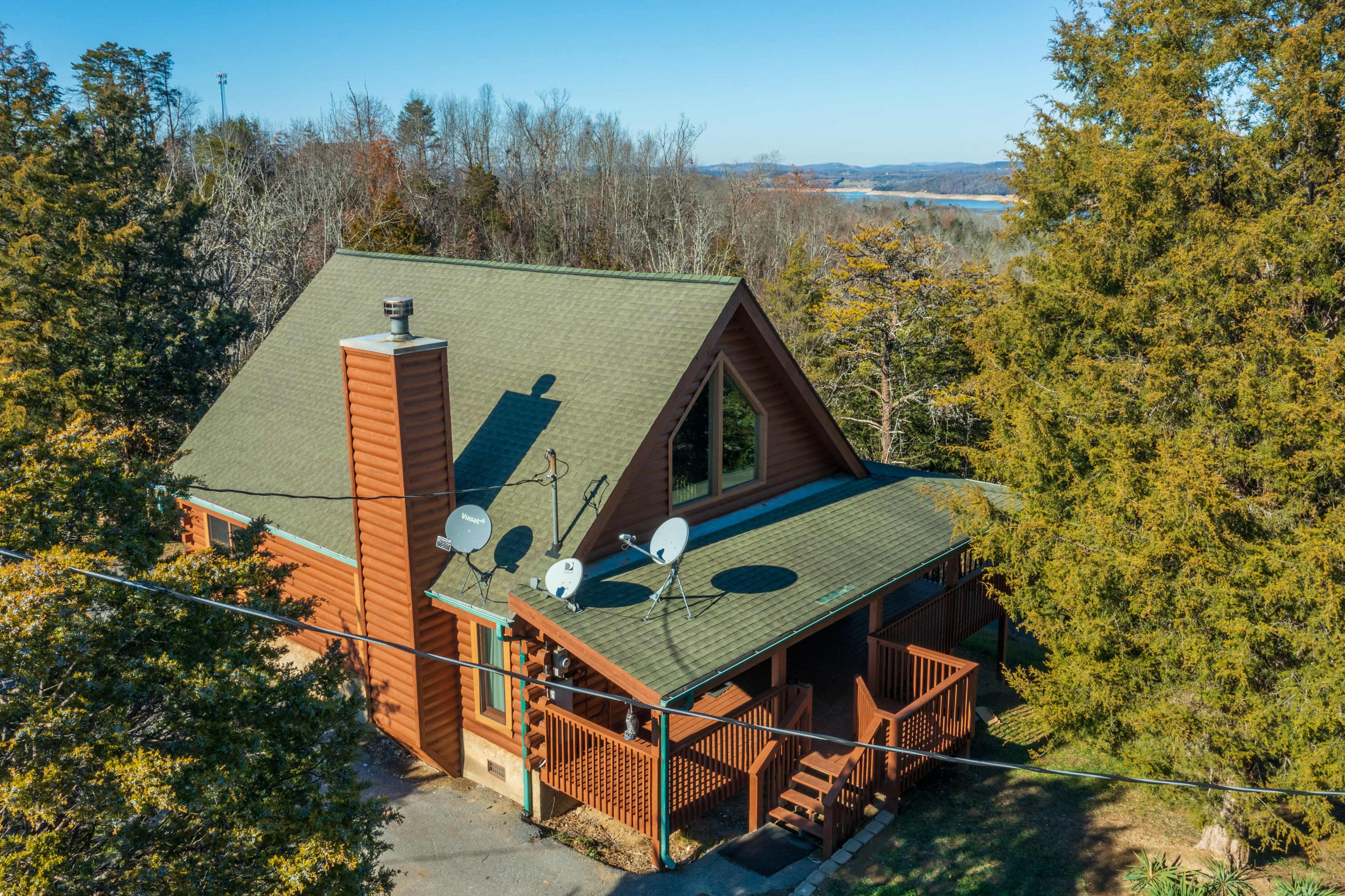 A wooden house with a sloped roof and satellite dishes sits surrounded by trees and overlooks a body of water in the distance.